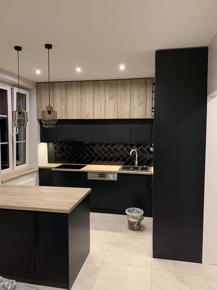 Nowoczesna kuchnia na wymiar: czarne szafki z drewnianymi akcentami, czarne płytki ułożone w jodełkę, wyspa kuchenna z blatem imitującym drewno i designerskie lampy.