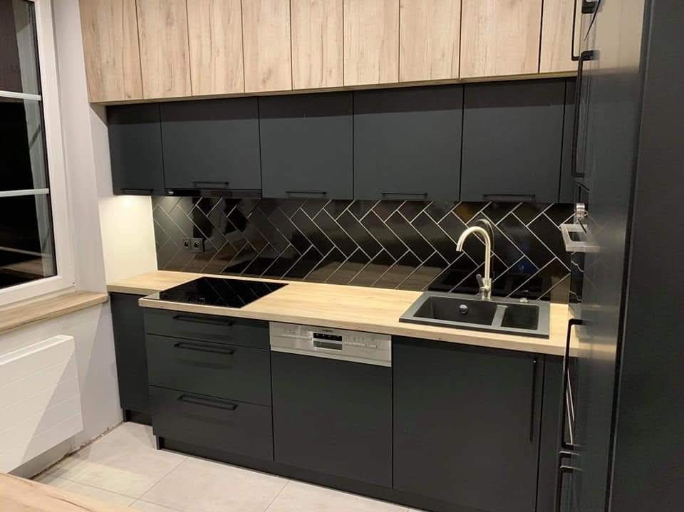 Nowoczesna kuchnia z czarnymi szafkami i blatem w odcieniu drewna. Uwagę przykuwają czarne płytki ułożone w jodełkę nad blatem. Minimalistyczny design, funkcjonalność i elegancja.
