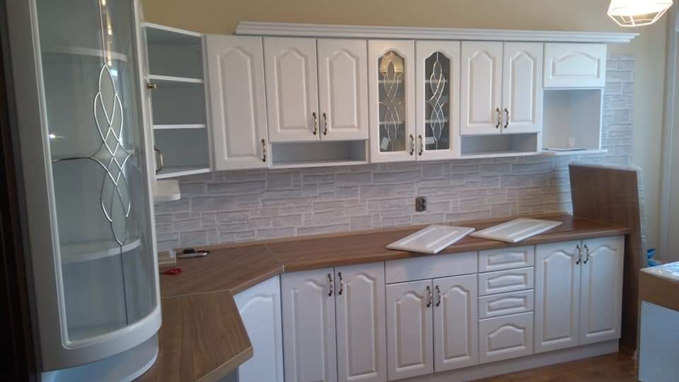 Biała, klasyczna zabudowa kuchenna z drewnianym blatem i szklanymi witrynami, wykończona dekoracyjną listwą. Widoczna część ściany z imitacją cegły.