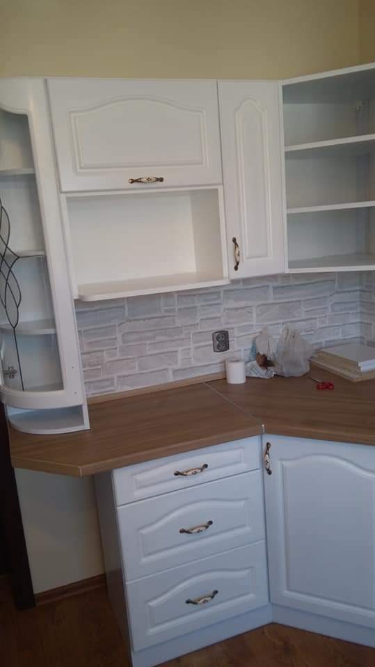 Biała, narożna zabudowa kuchenna z drewnianym blatem i ozdobnymi uchwytami, na tle ściany z cegieł. Widoczne szafki górne i dolne, z otwartymi półkami.