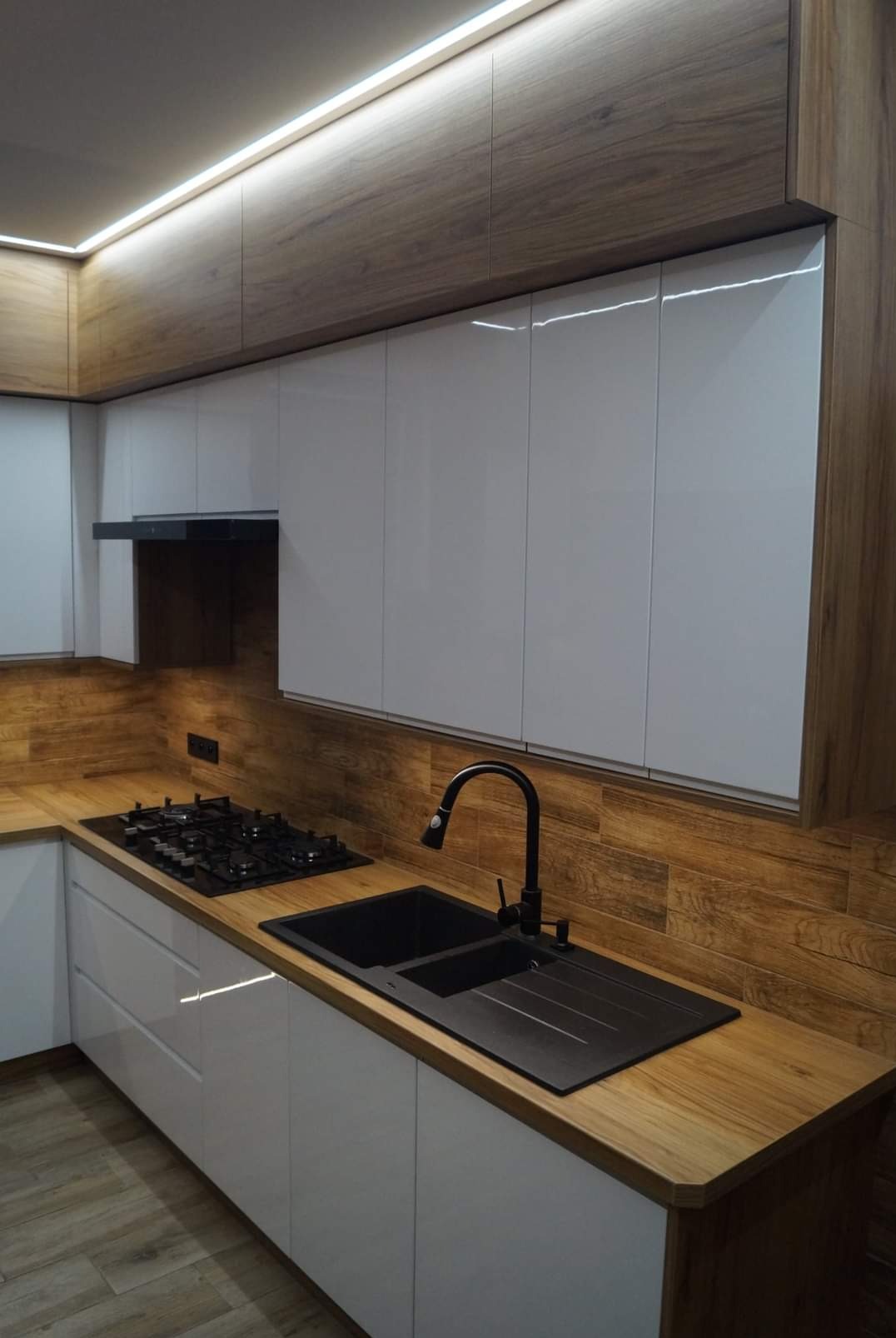 Nowoczesna kuchnia z białymi, lakierowanymi frontami, drewnianym blatem i oświetleniem LED. Czarny zlew i płyta gazowa dodają elegancji. Widok na zabudowę kuchenną.