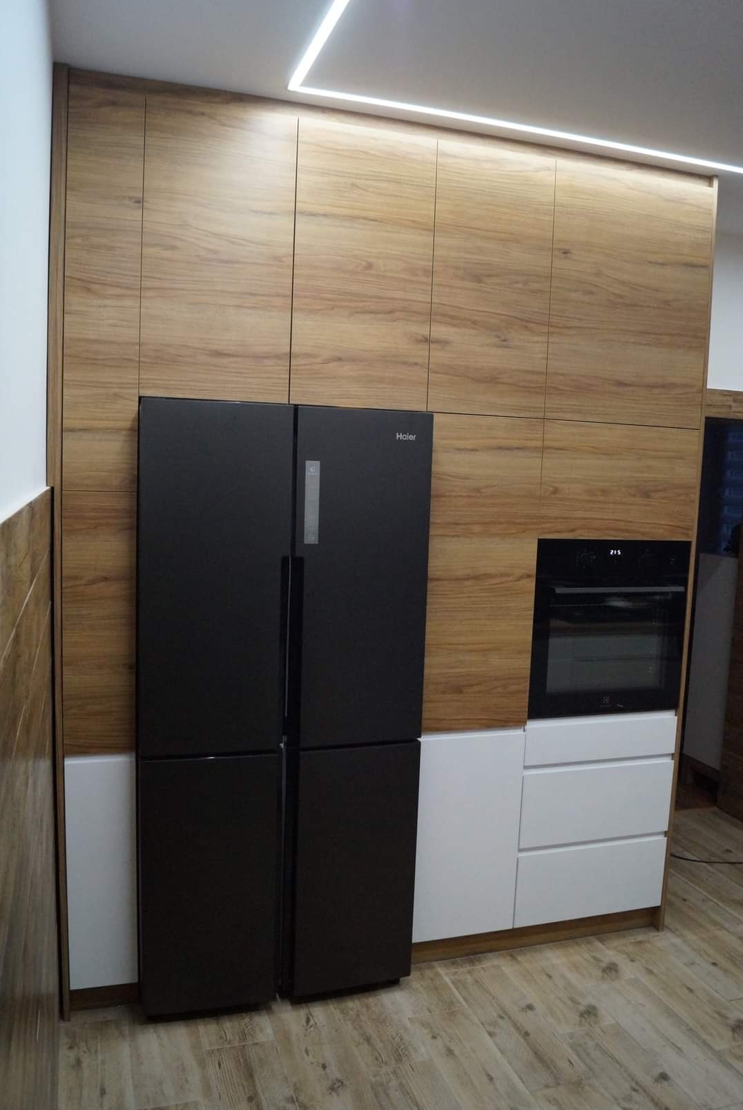 Nowoczesna kuchnia z drewnianymi frontami szafek, czarną lodówką side-by-side i piekarnikiem w zabudowie. Białe dolne szafki i oświetlenie LED pod sufitem.