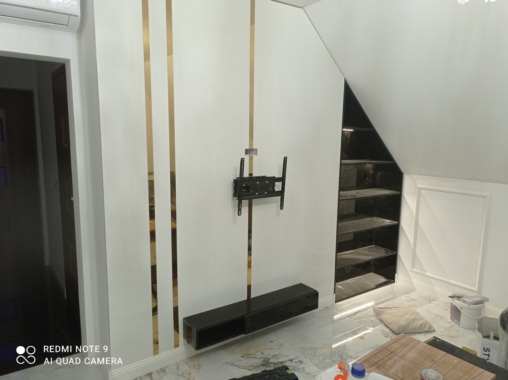 Biała ściana z dekoracyjnymi, złotymi paskami, czarna szafka RTV i uchwyt na telewizor. W tle widoczna czarna zabudowa wnęki z półkami i marmurowa podłoga.