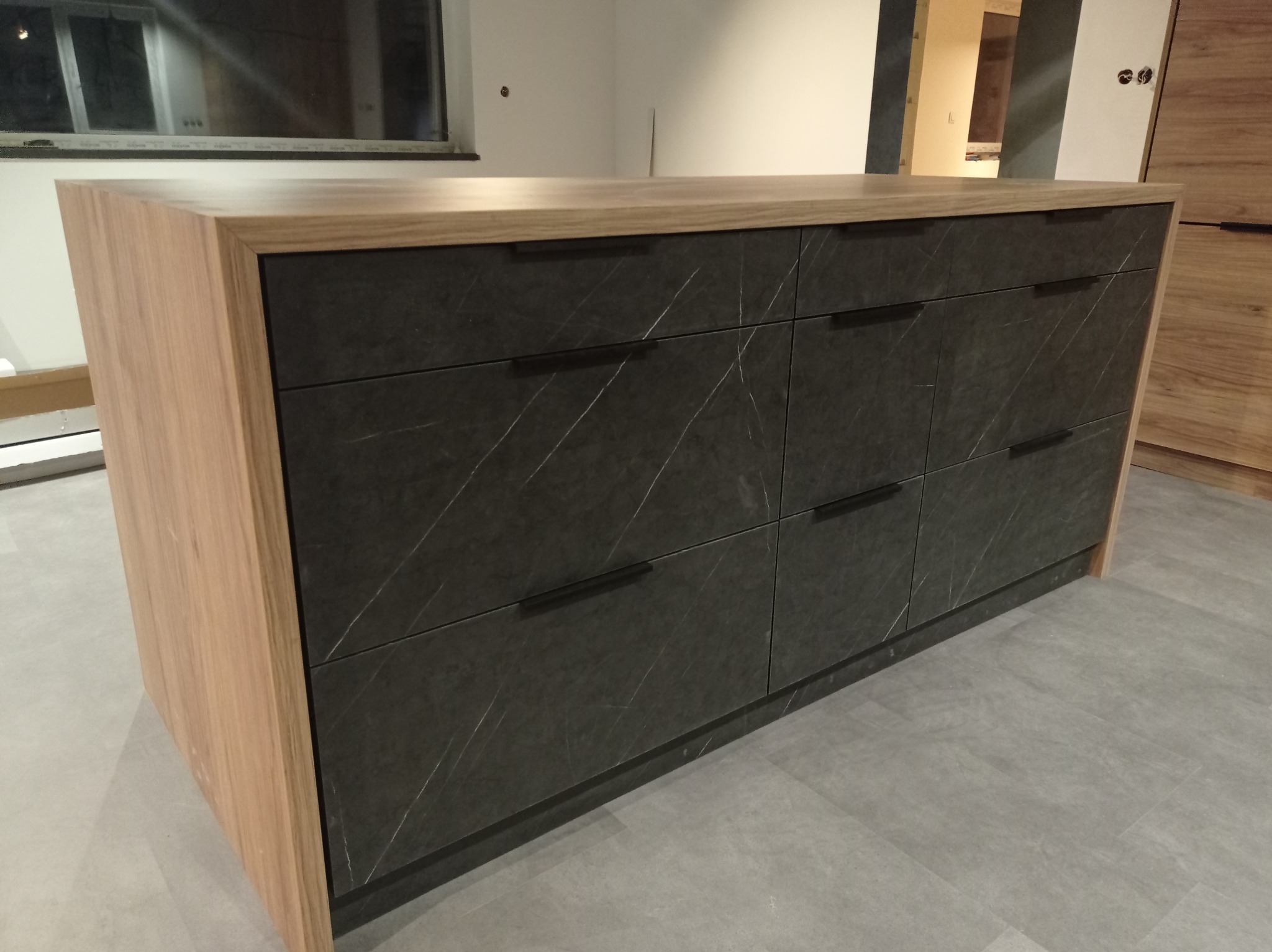 Wyspa kuchenna z blatem z jasnego drewna i frontami szuflad w kolorze grafitowym z marmurkowym wzorem. Minimalistyczne, czarne uchwyty. Szara podłoga.