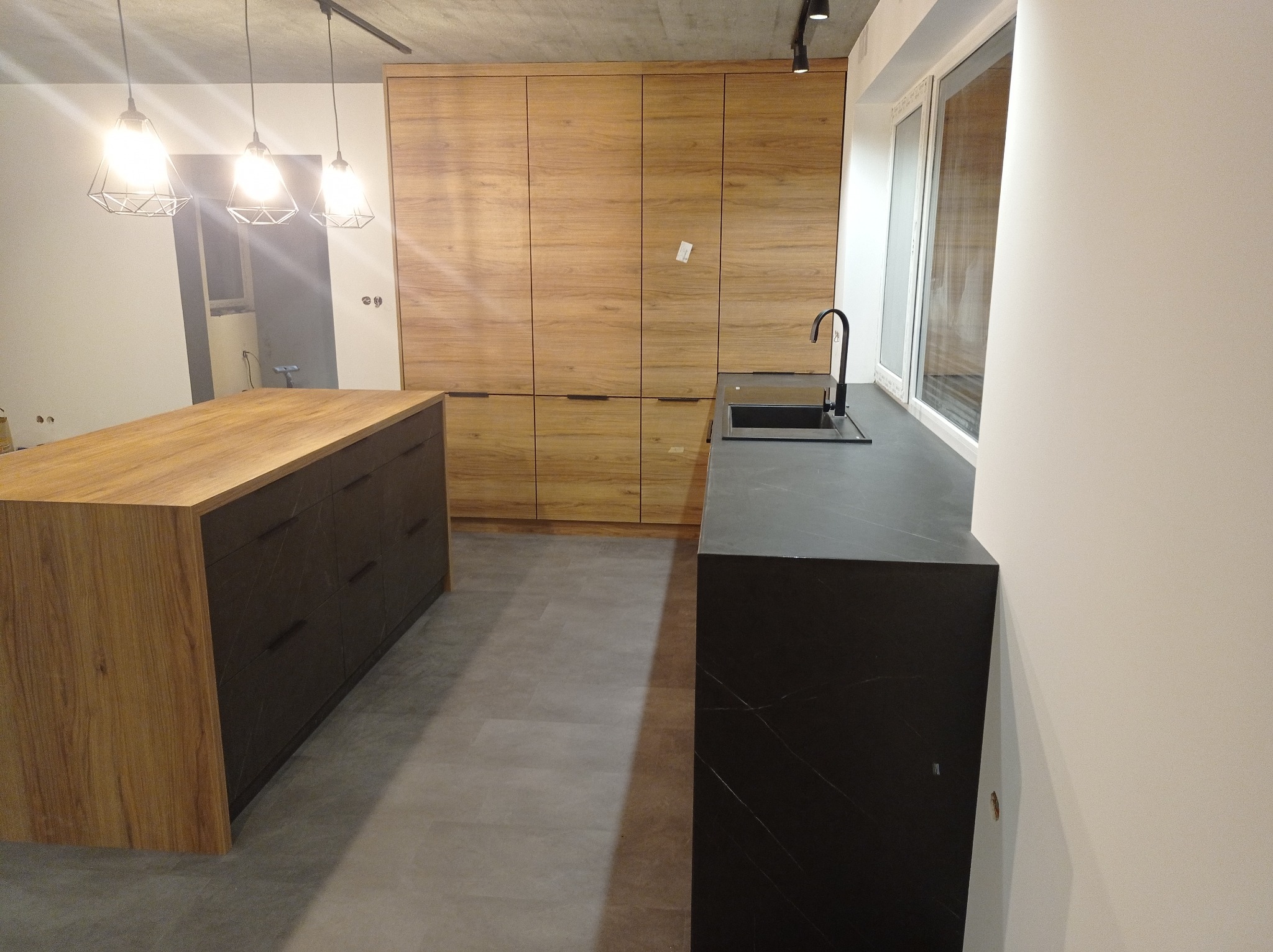 Nowoczesna kuchnia z wyspą i zabudową drewnianą pod sufit. Czarny blat i minimalistyczna armatura. Oświetlenie punktowe i duże okno doświetlają wnętrze.