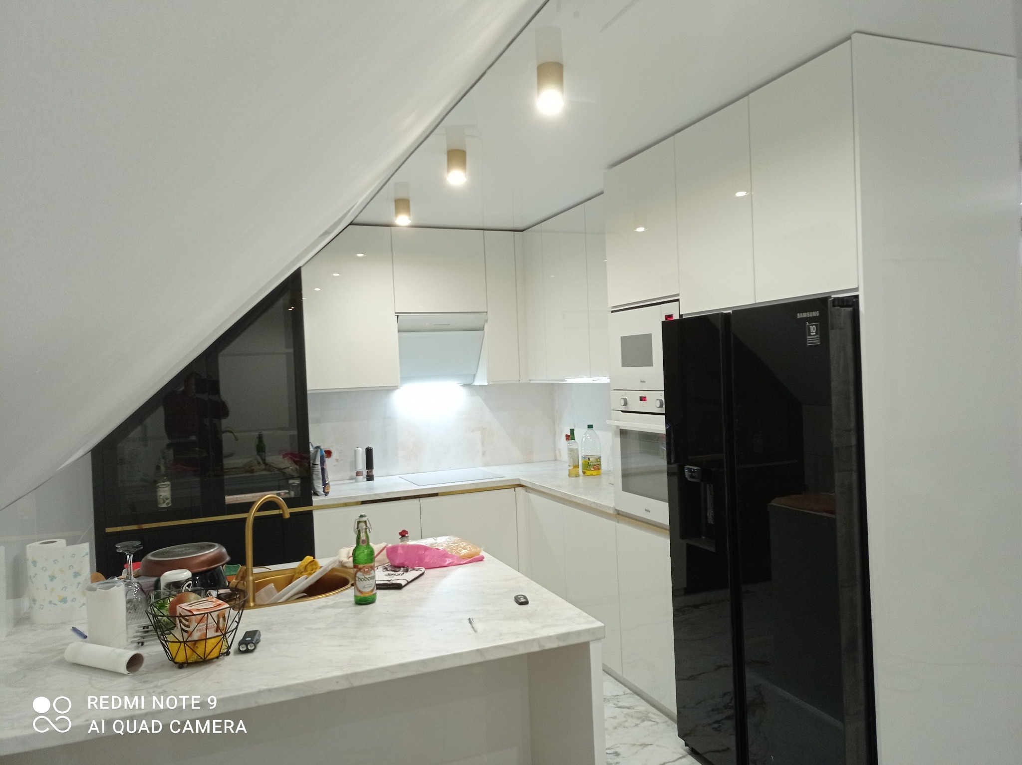 Biała kuchnia z połyskiem w skosach poddasza, oświetlenie punktowe, marmurowy blat, czarna lodówka. Widok na zlewozmywak i okno. Nowoczesny design.