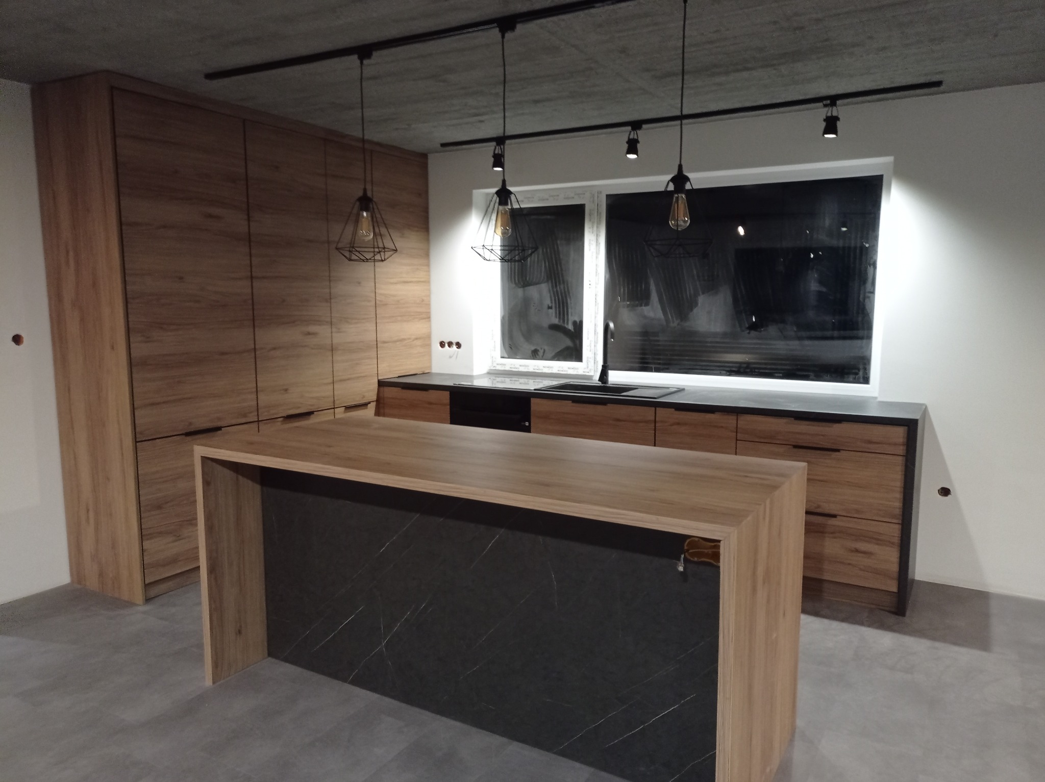 Nowoczesna kuchnia z wyspą i drewnianymi frontami, oświetlona lampami wiszącymi. Minimalistyczny design, szare płytki podłogowe, okno z widokiem na zewnątrz.