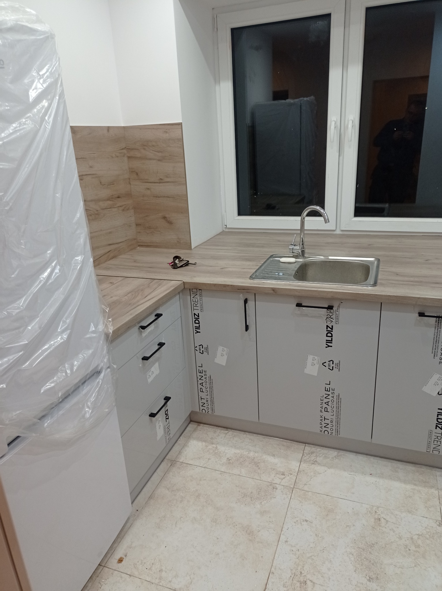 Jasna kuchnia z drewnianym blatem i szarymi frontami szafek, widoczny zlew i bateria. Częściowo widoczna lodówka w folii ochronnej. Ułożone płytki podłogowe.