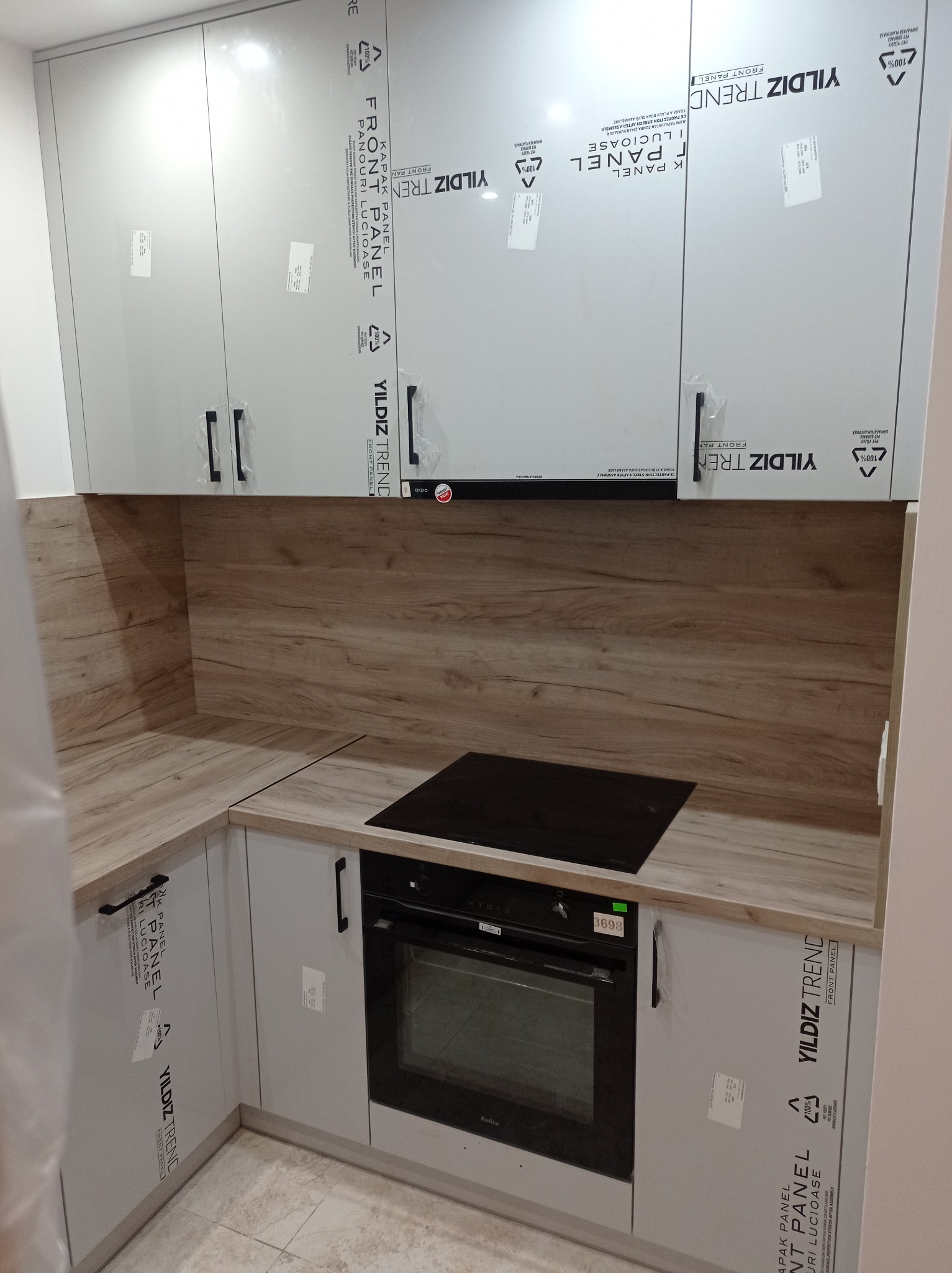 Nowoczesna kuchnia z szarymi szafkami, drewnianym blatem i czarną płytą indukcyjną. Fronty szafek zabezpieczone folią ochronną. Ujęcie z perspektywy podłogi.