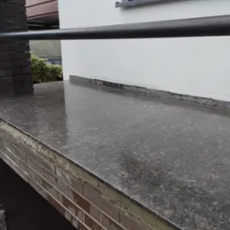 Zewnętrzny taras z ciemnego, polerowanego kamienia z ceglaną konstrukcją nośną i czarną metalową barierką, lekko mokry od deszczu.