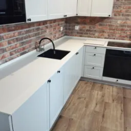 Kremowy blat kuchenny z czarnym zlewem i baterią, białe szafki z czarnymi gałkami, ściana z cegły, drewnopodobne płytki podłogowe, płyta indukcyjna.