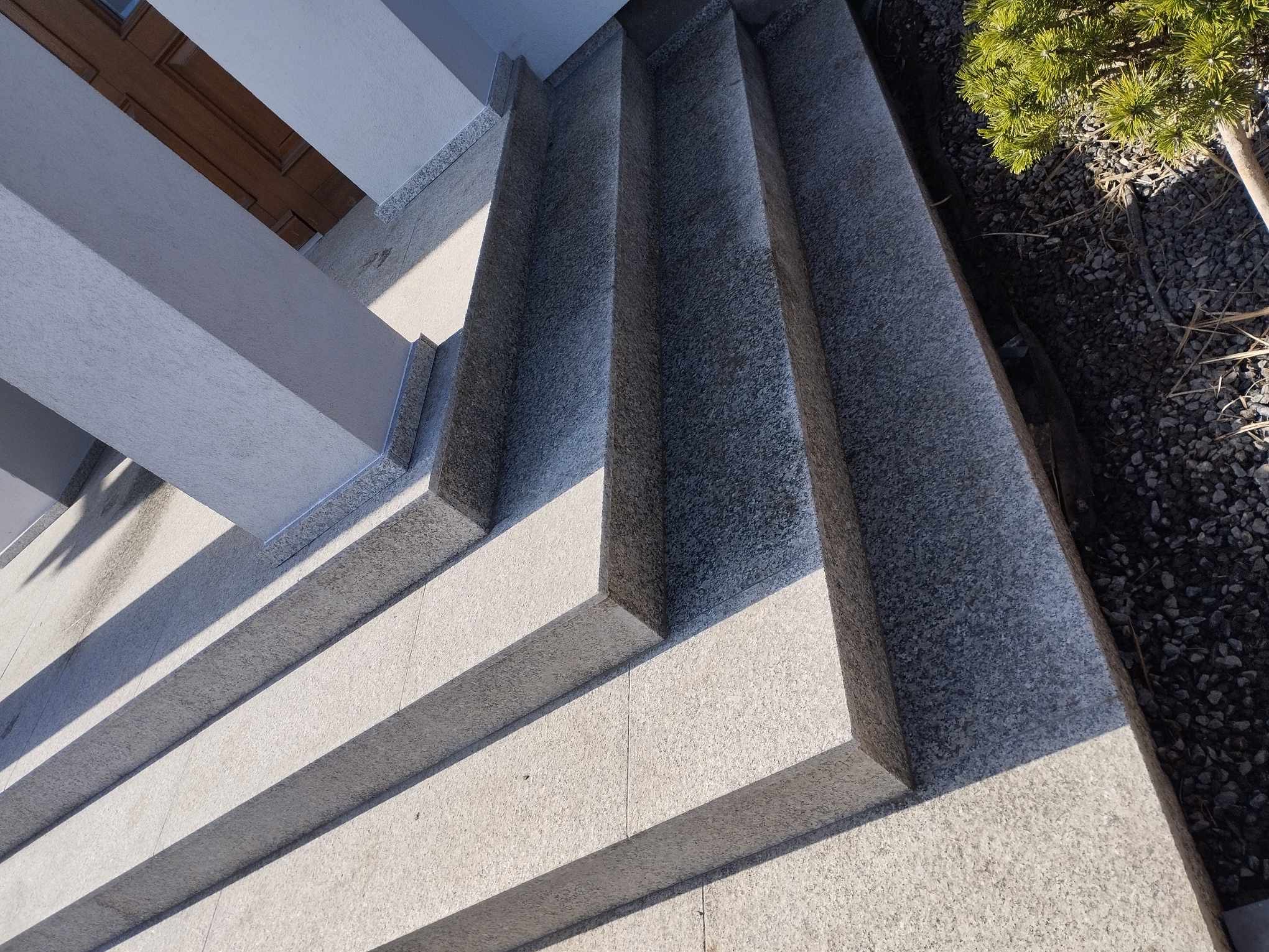 Granitowe schody zewnętrzne prowadzące do drzwi wejściowych, z widocznym fragmentem kolumny i roślinności obok.