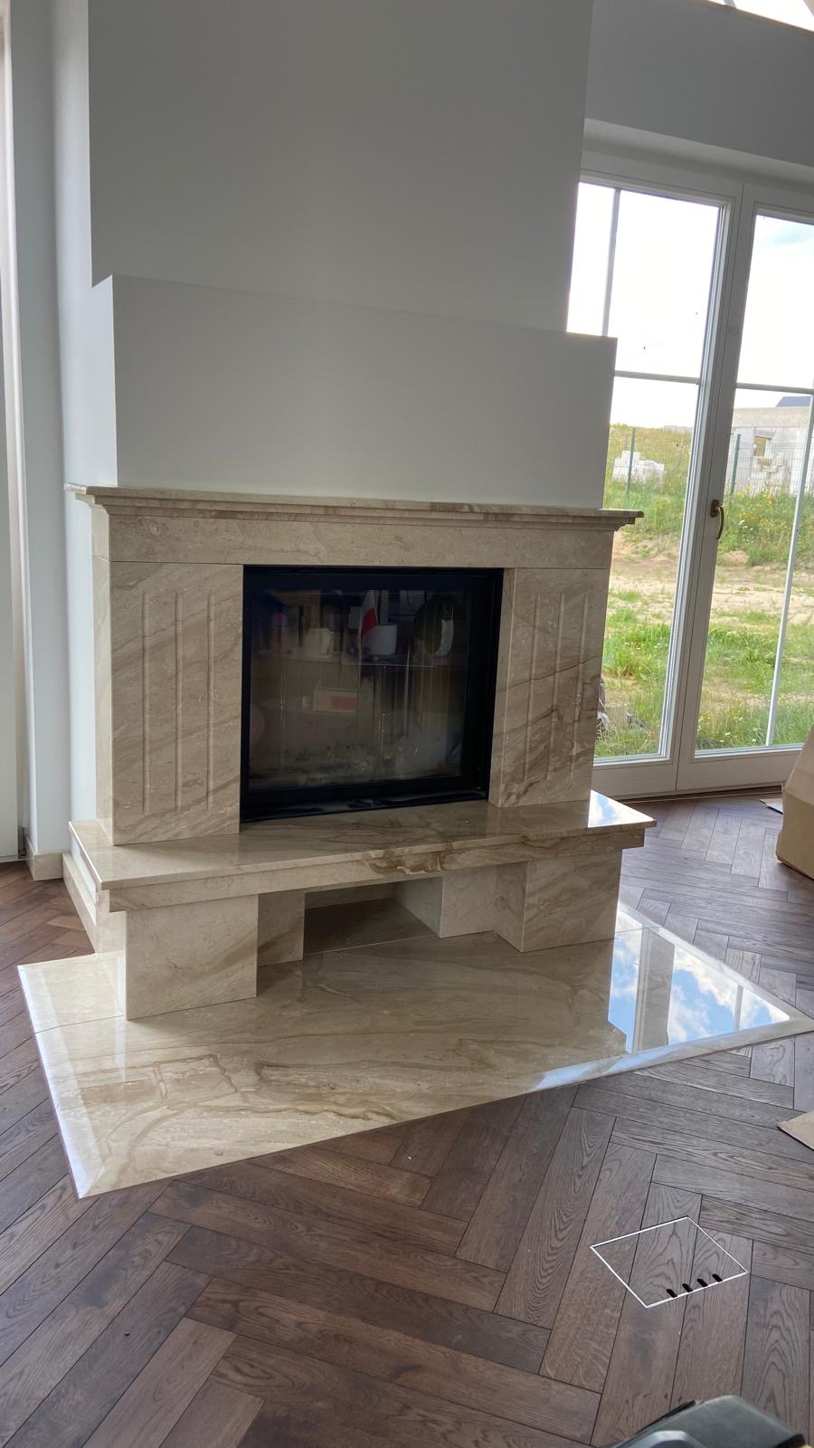 Marmurowy kominek z czarnym wkładem, z widocznym odbiciem nieba w polerowanej płycie podłogowej, ułożonej na parkiecie w jodełkę.