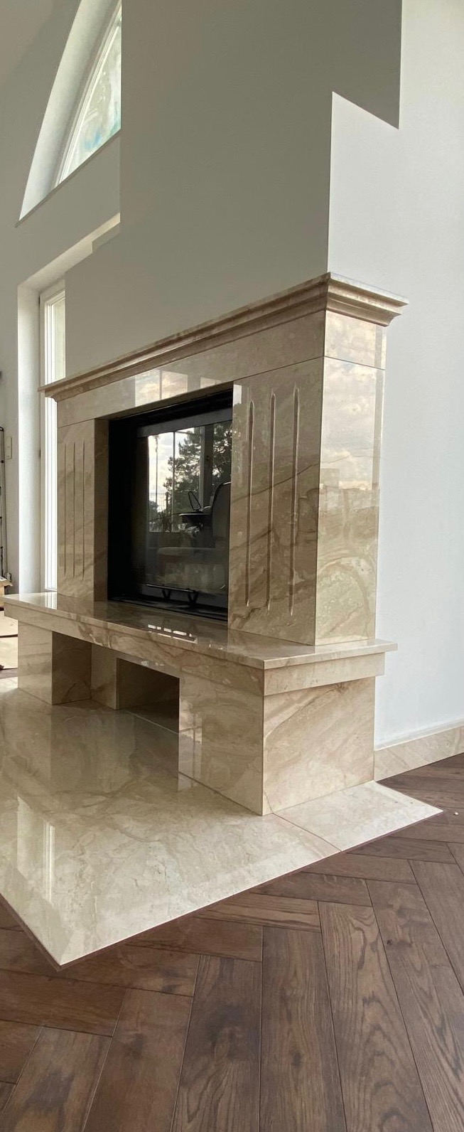 Obudowa kominkowa z beżowego kamienia z wyraźnym żyłkowaniem, z wkładem kominkowym, na podłodze z płytek kamiennych przechodzących w parkiet ułożony w jodełkę.
