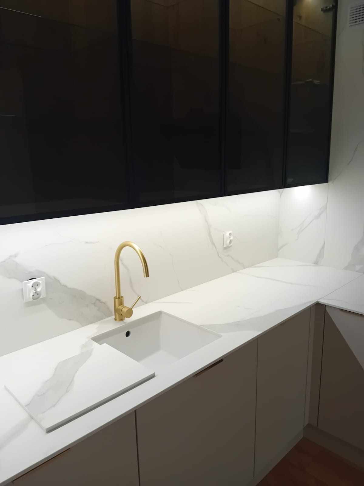 Biały blat kuchenny z marmurowym wzorem, złota bateria przy białym zlewozmywaku, czarne szafki wiszące i podświetlenie LED pod szafkami.