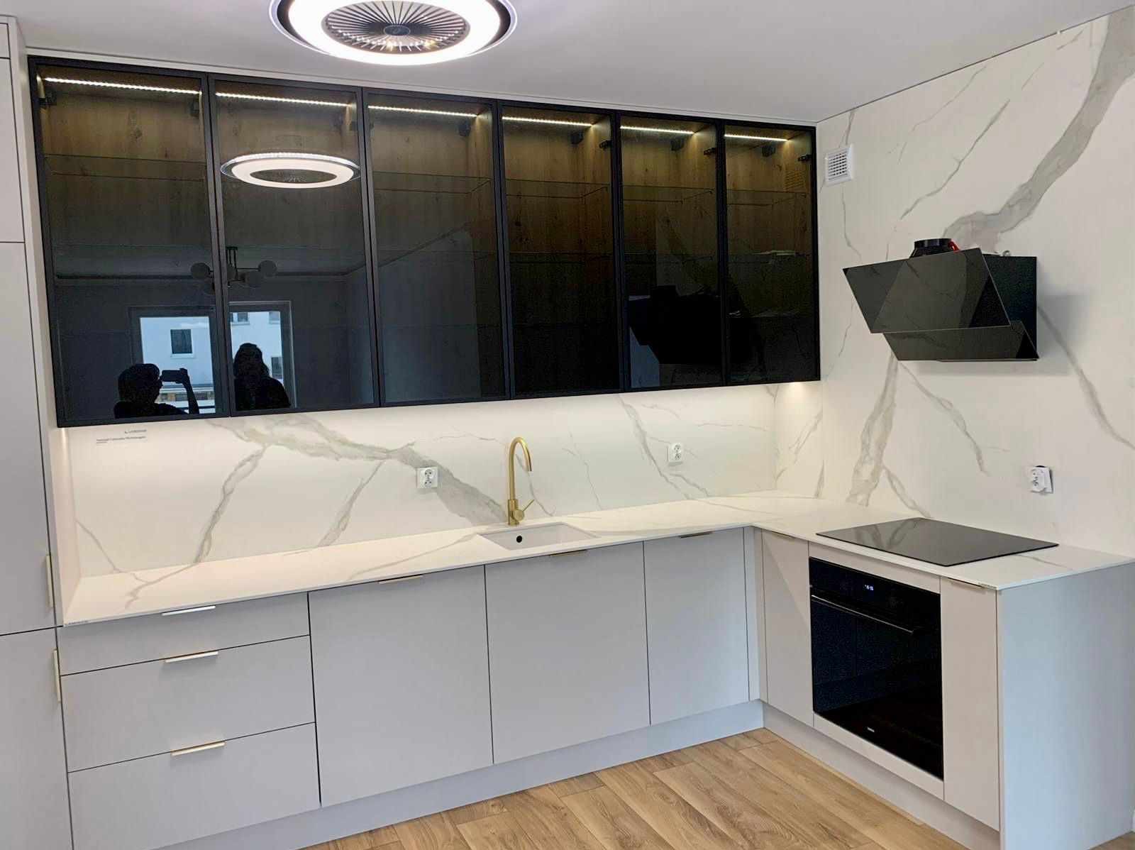 Nowoczesna kuchnia z białymi szafkami, blatem imitującym marmur, złotą armaturą i czarnym okapem, oświetlona lampami sufitowymi