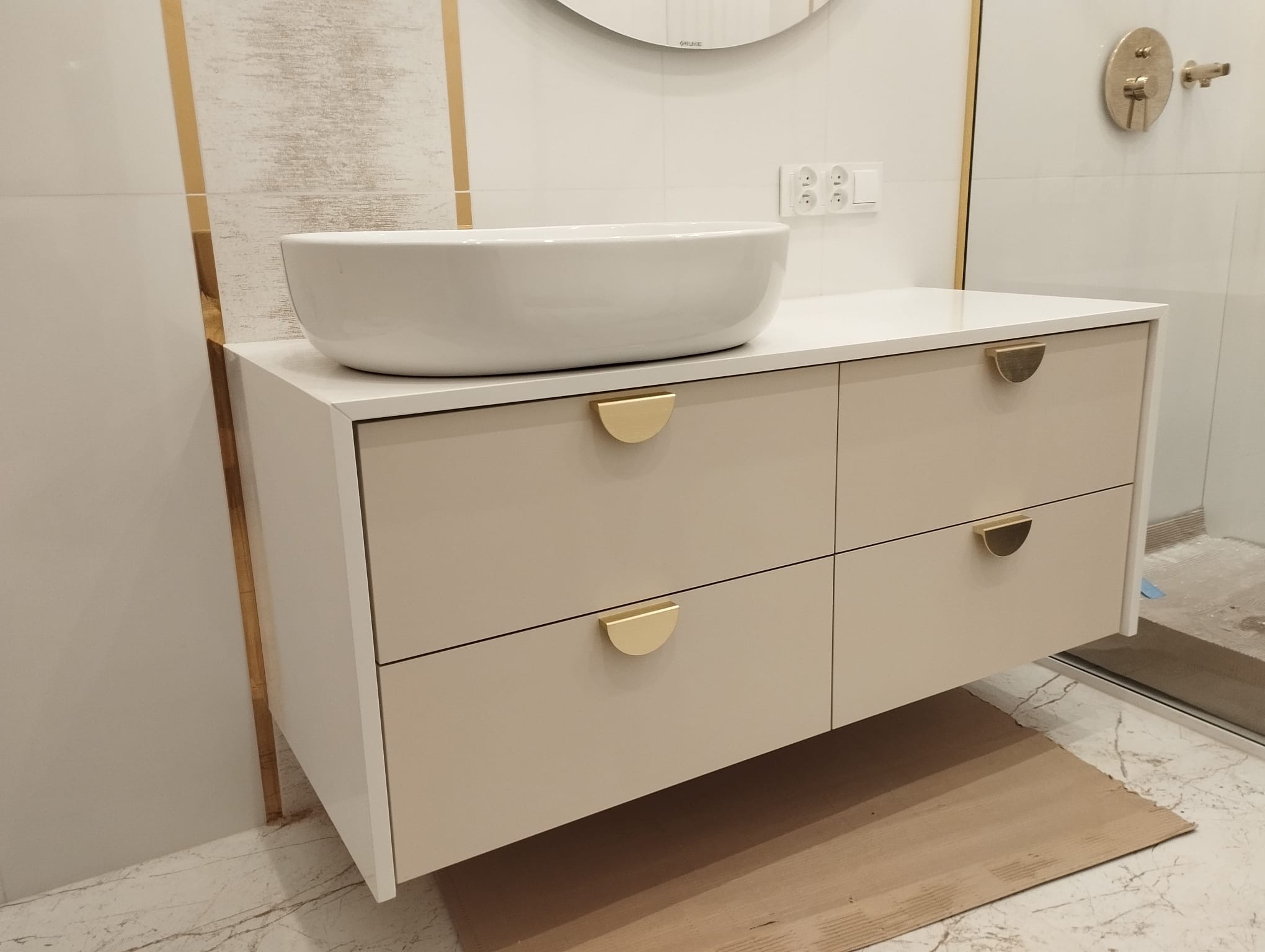 Biała umywalka nablatowa na szafce z beżowymi frontami szuflad i półokrągłymi, złotymi uchwytami, na marmurowej posadzce z kartonem ochronnym, widoczne złote akcenty na ścianie i armatura.