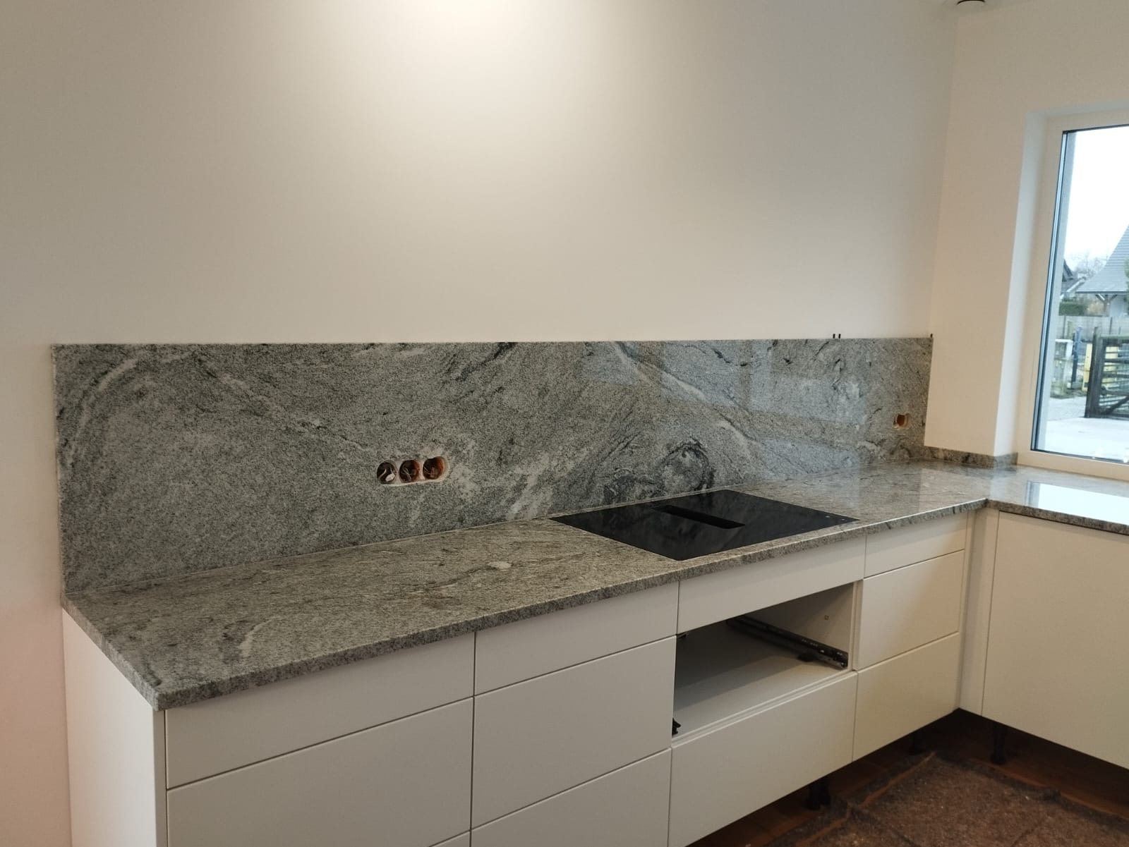 Kuchnia w trakcie wykańczania: szary blat z kamienia naturalnego z zamontowaną płytą indukcyjną i wyciętymi otworami na gniazdka elektryczne, białe fronty szafek kuchennych, widok na okno.