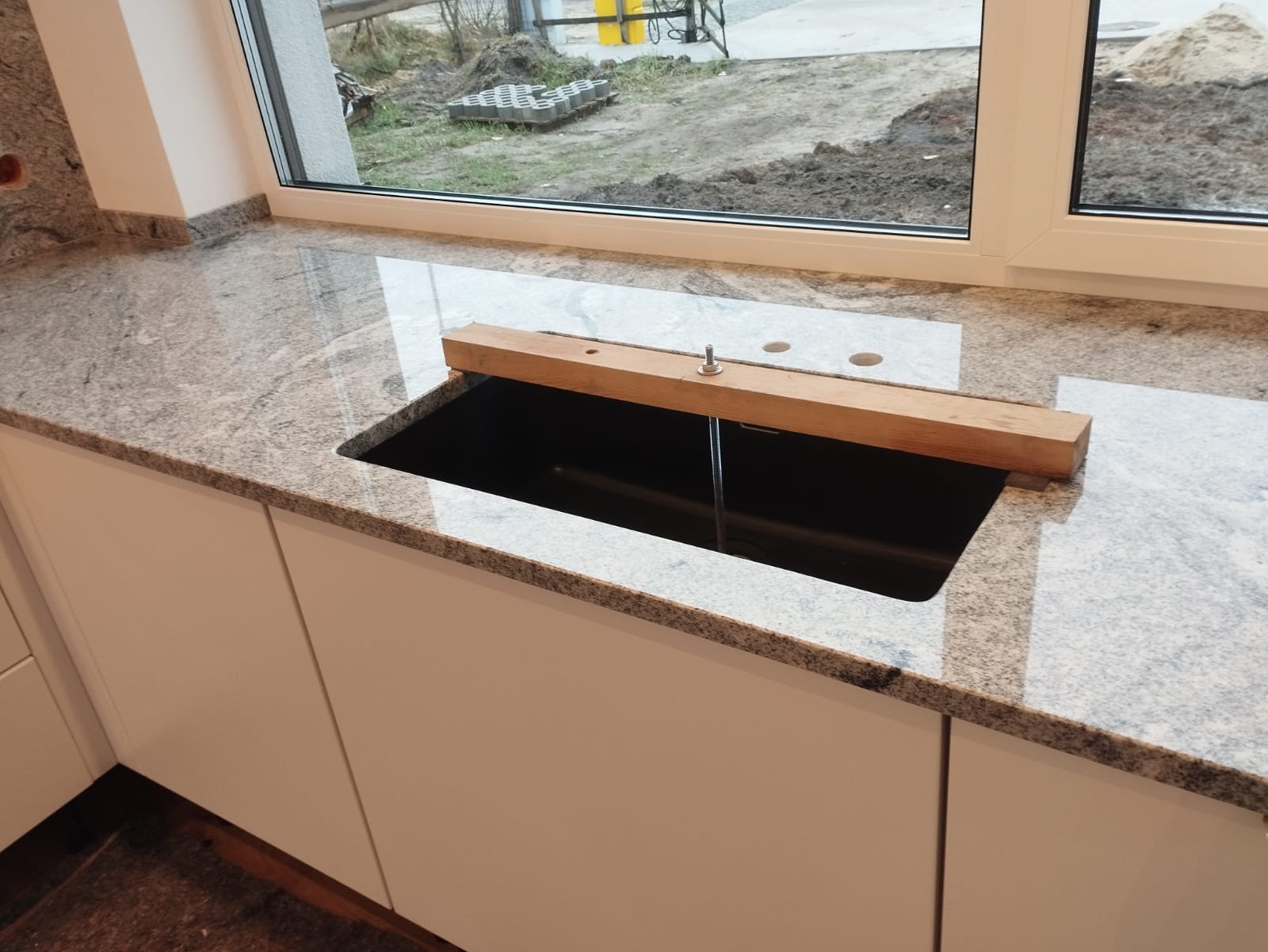 Montaż zlewu w blacie z jasnego granitu przy oknie, drewniana belka podtrzymująca zlew, widoczne otwory na baterię.