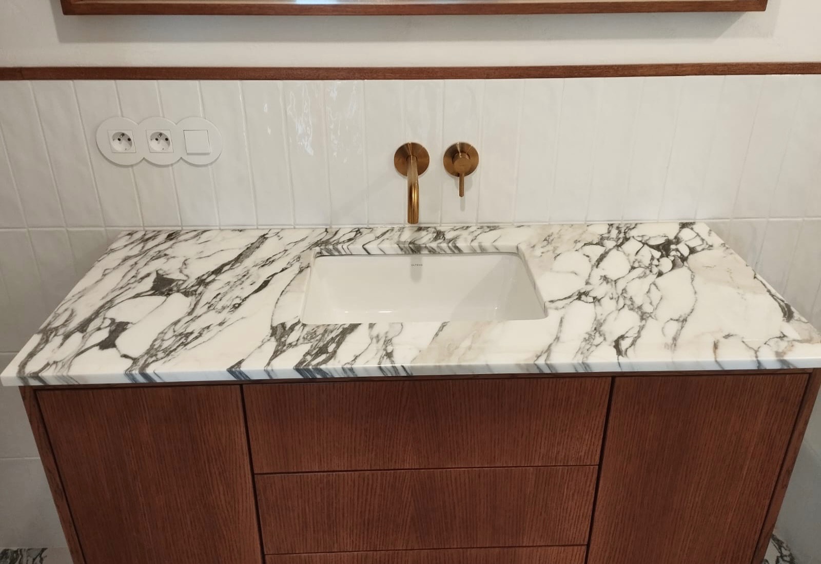 Łazienka z blatem z białego marmuru z szarymi żyłami, umywalką wpuszczaną i złotą armaturą na tle białych płytek.