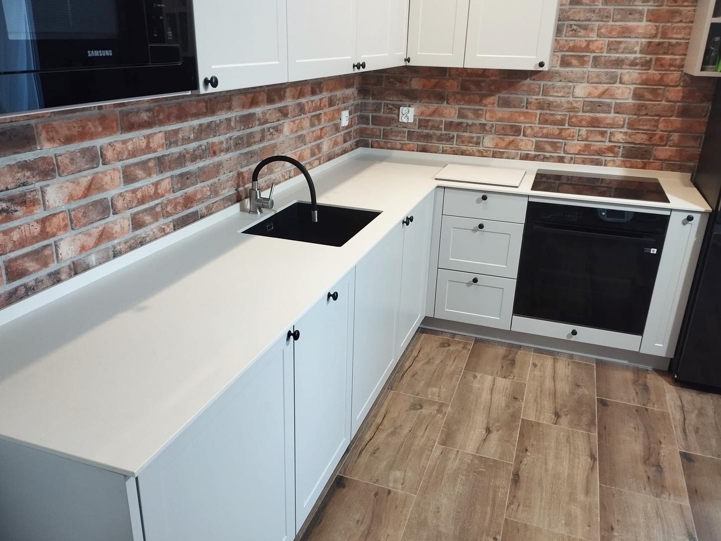 Kremowy blat kuchenny z czarnym zlewem i baterią, białe szafki z czarnymi gałkami, ściana z cegły, drewnopodobne płytki podłogowe, płyta indukcyjna.