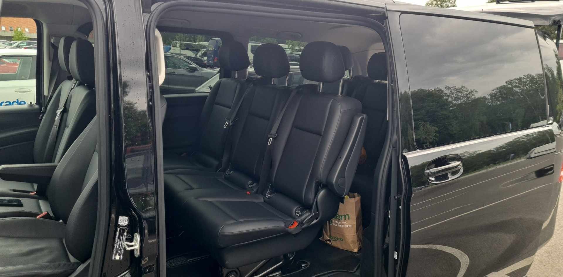 Wnętrze czarnego busa z trzema rzędami skórzanych foteli, widok z otwartych drzwi bocznych, papierowa torba na podłodze.