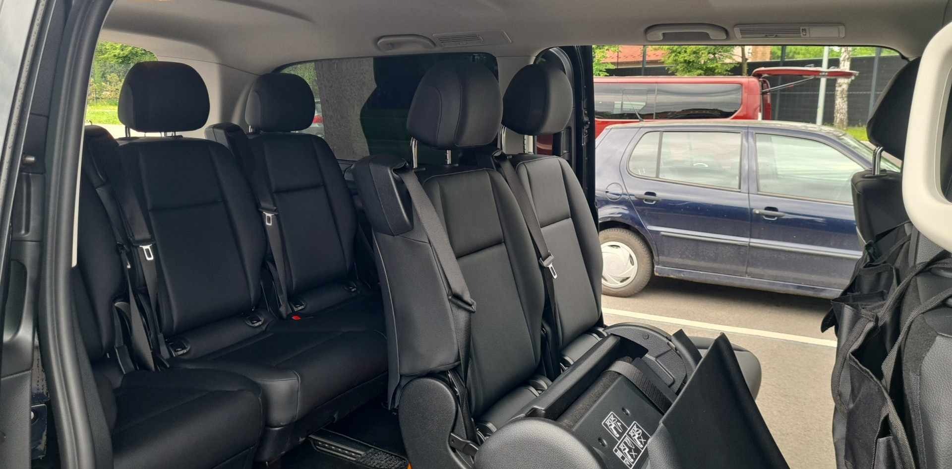 Wnętrze przestronnego busa z czarnymi skórzanymi siedzeniami i rozkładanym stolikiem, widok z perspektywy pasażera na zaparkowane na zewnątrz samochody.