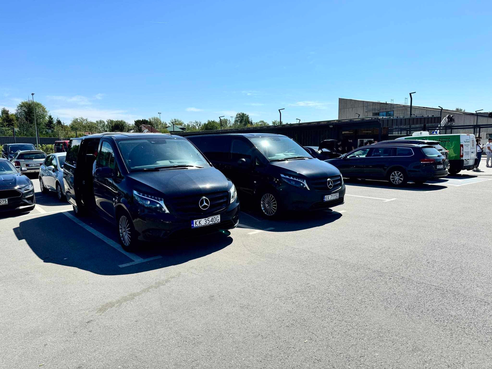 Dwa czarne busy Mercedes-Benz zaparkowane na parkingu w słoneczny dzień, z otwartymi drzwiami jednego z nich, widoczne tablice rejestracyjne.