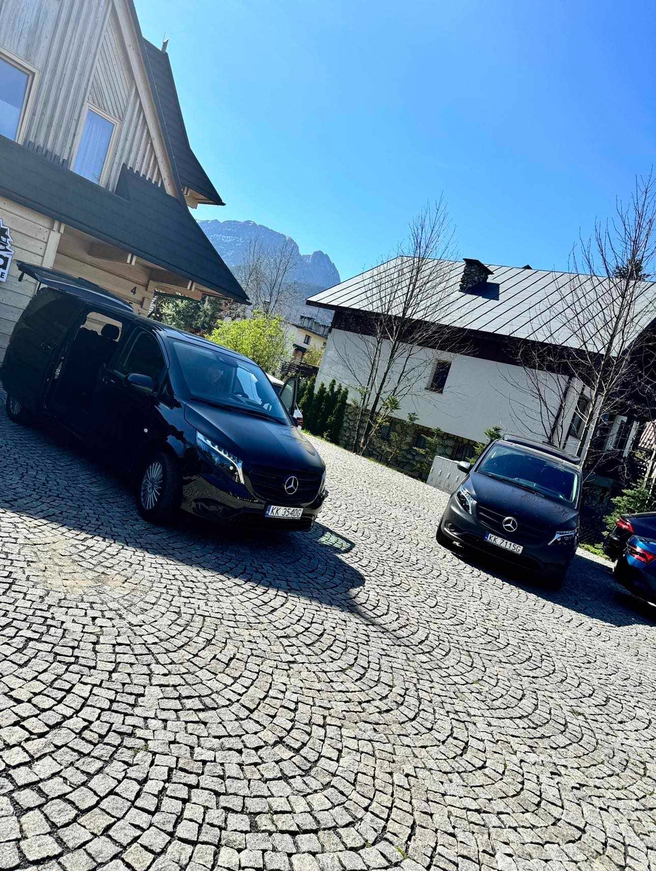 Dwa czarne busy Mercedes Vito zaparkowane na brukowanej posesji przed domami w stylu góralskim, widok na góry w tle, słoneczny dzień.