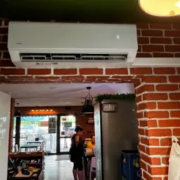 Biała klimatyzacja AUX zamontowana na ścianie z czerwonej cegły w lokalu usługowym, widok z dołu na wnętrze lokalu z klientami.
