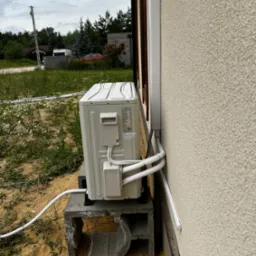 Zewnętrzna jednostka klimatyzacji zamontowana na betonowym fundamencie przy ścianie domu z elewacją typu baranek, widoczne rury odprowadzające czynniki chłodnicze w białej osłonie.