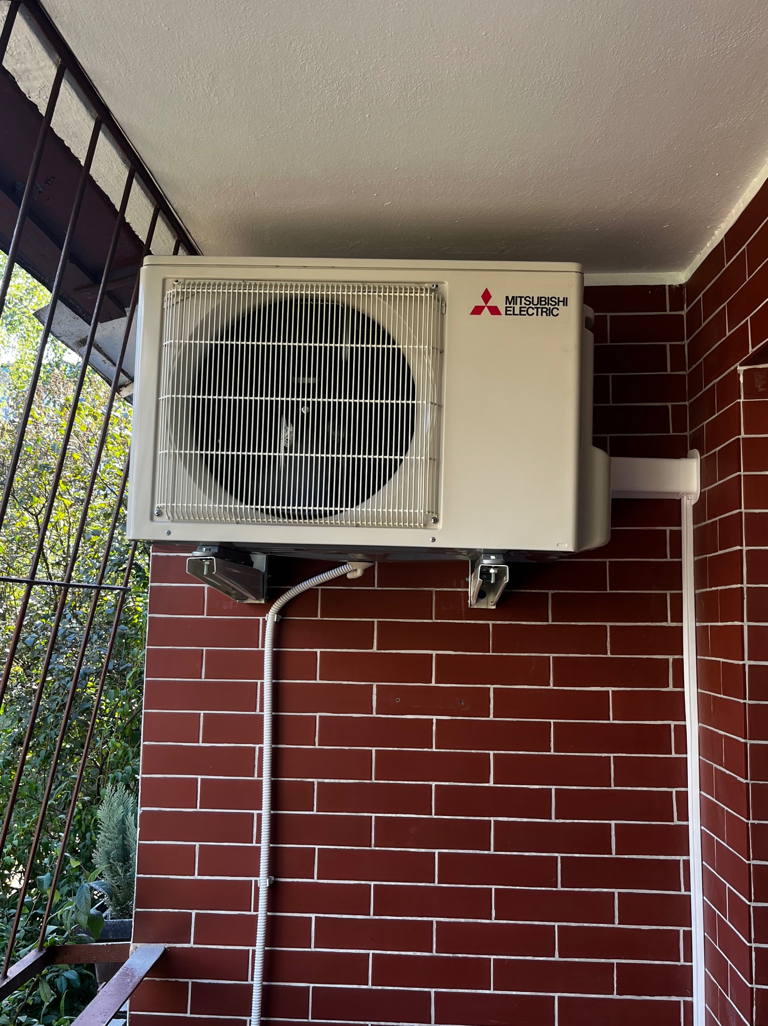 Zewnętrzna jednostka klimatyzacji Mitsubishi Electric zamontowana na ceglanej ścianie balkonu, widoczne przewody i elementy montażowe.