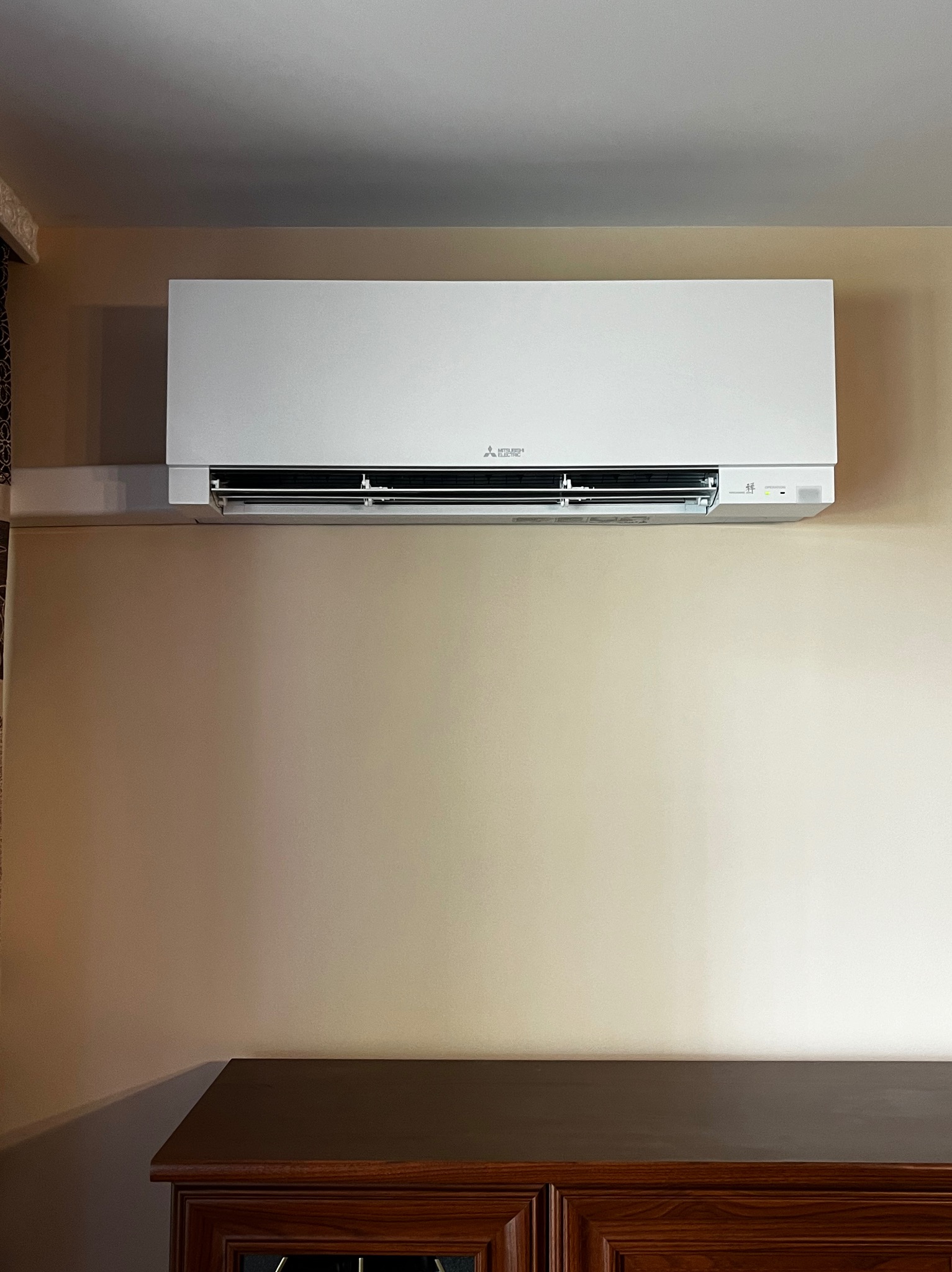Biały klimatyzator Mitsubishi Electric zamontowany na beżowej ścianie w pomieszczeniu, widoczny fragment ciemnobrązowej komody poniżej.