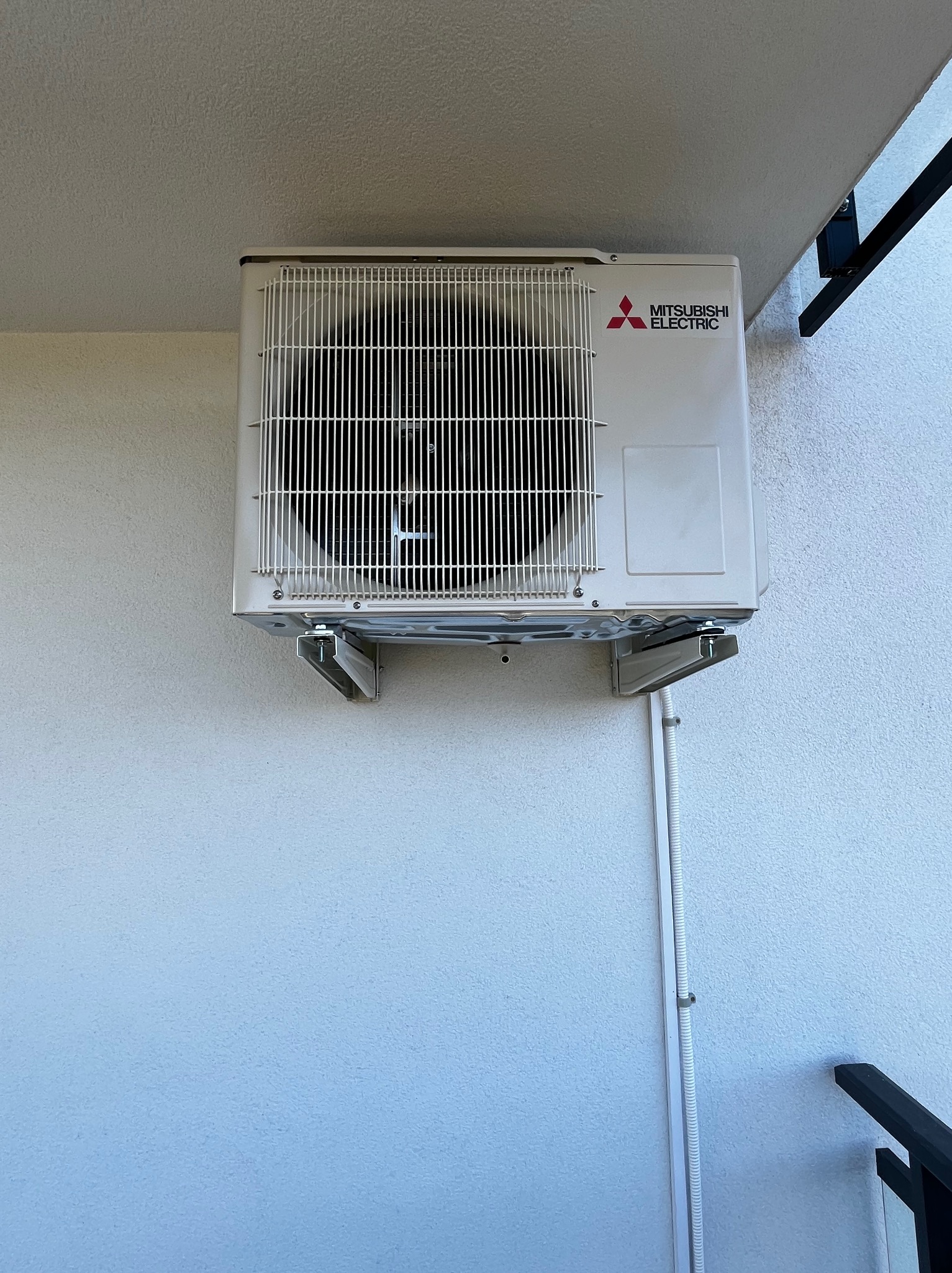 Zewnętrzna jednostka klimatyzatora Mitsubishi Electric zamontowana na jasnej elewacji budynku, widoczne przewody w białej listwie. Fragment balustrady w rogu kadru.