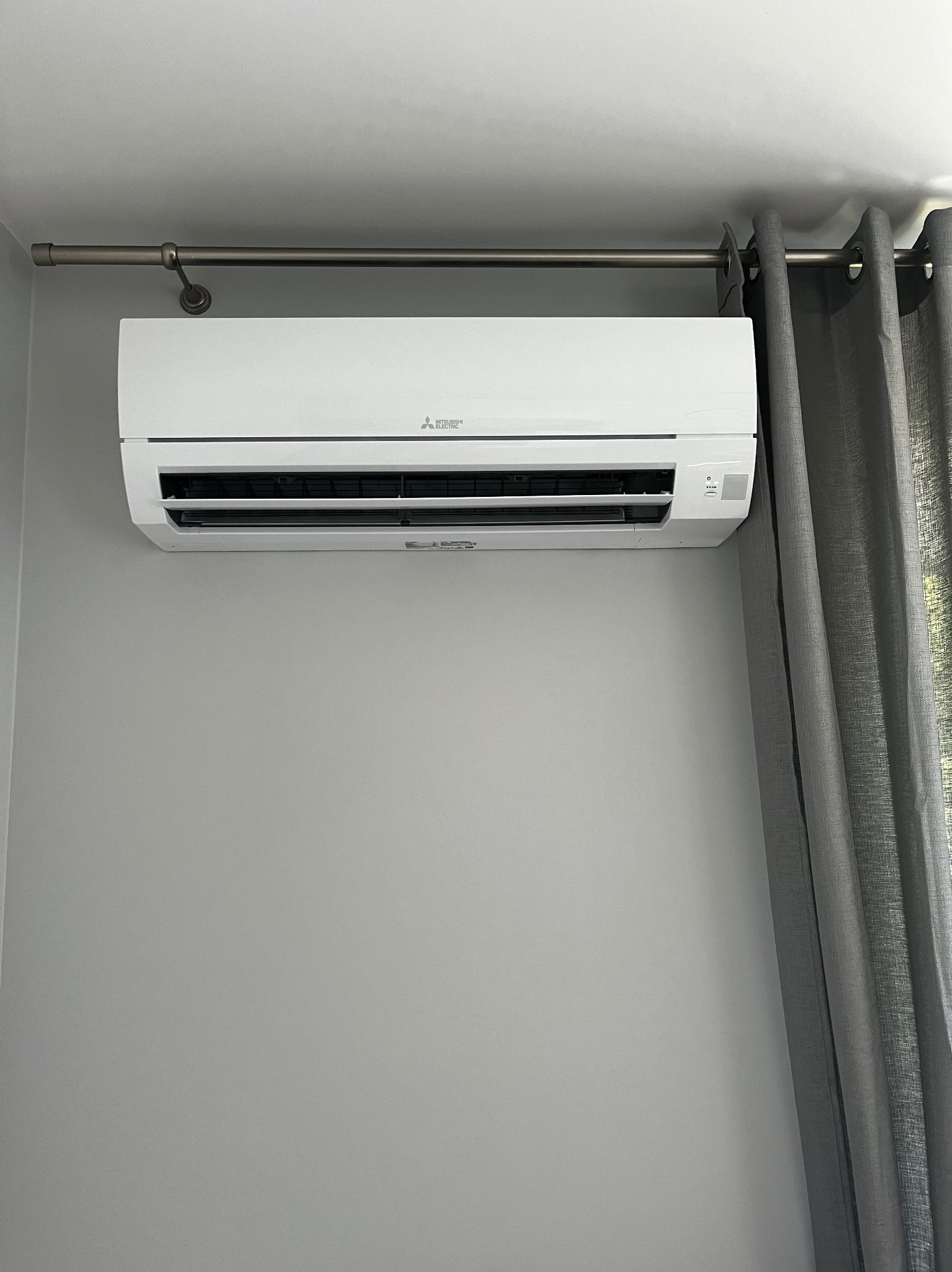 Biała jednostka klimatyzacji Mitsubishi Electric zamontowana na szarej ścianie, widoczne szare zasłony po prawej stronie i karnisz nad urządzeniem.