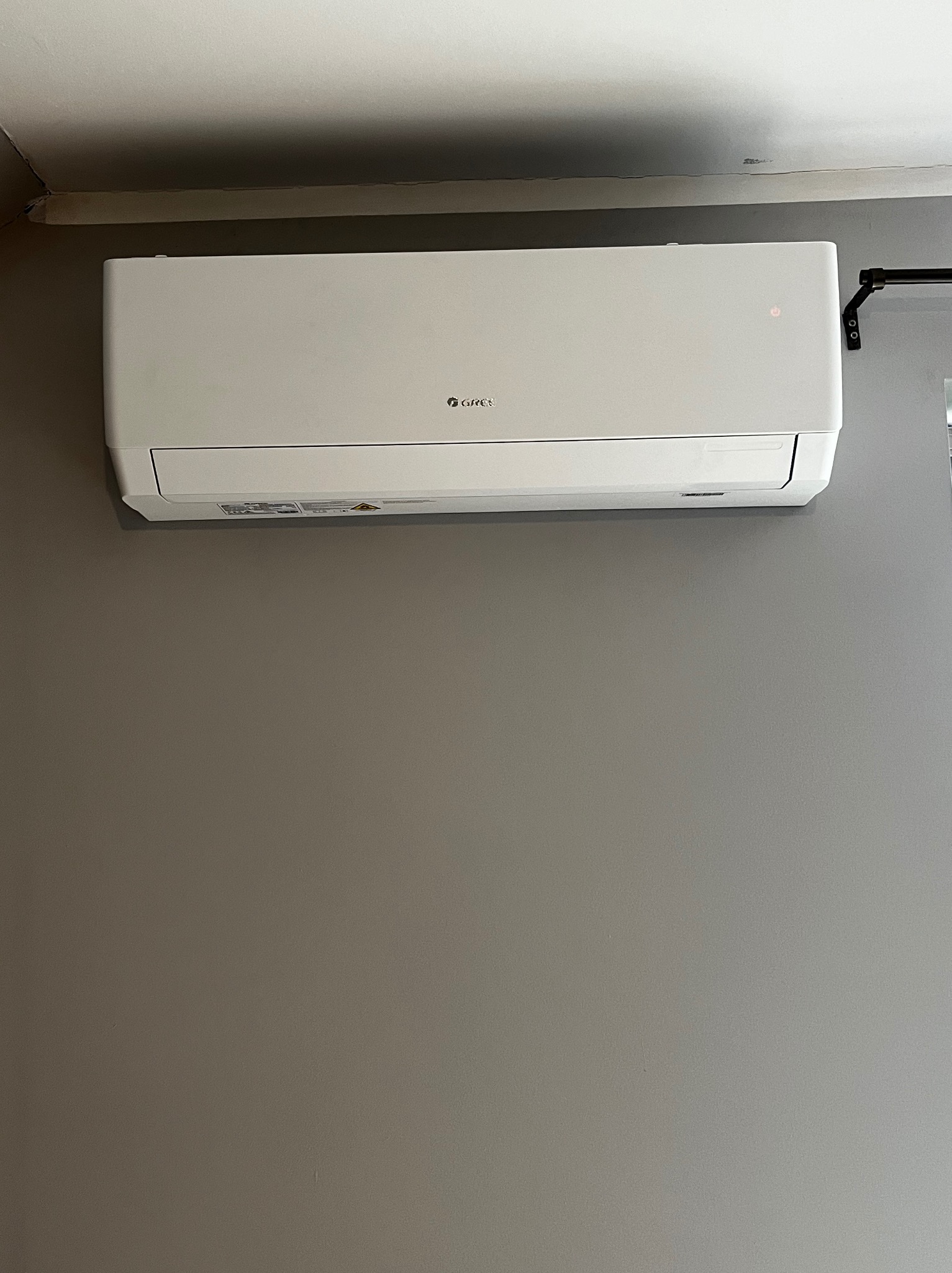 Biały klimatyzator marki Gree zamontowany na szarej ścianie w pomieszczeniu, widoczny fragment sufitu i karnisza.