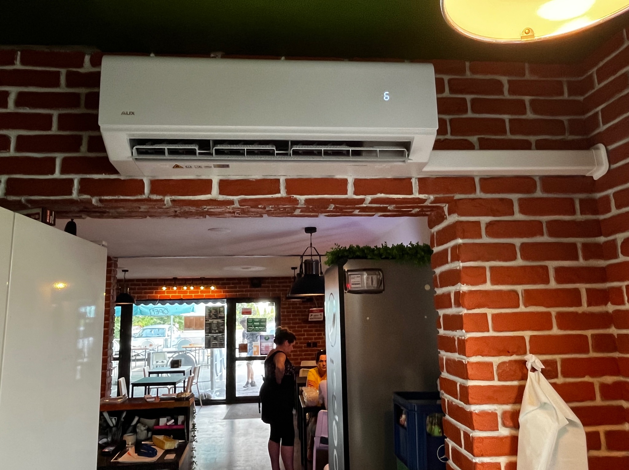 Biała klimatyzacja AUX zamontowana na ścianie z czerwonej cegły w lokalu usługowym, widok z dołu na wnętrze lokalu z klientami.