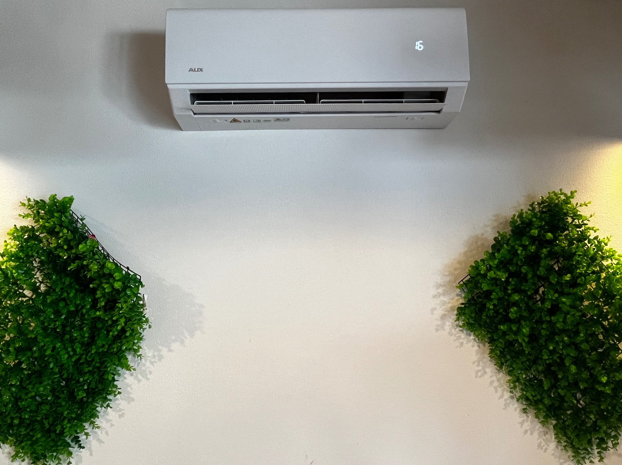 Biały klimatyzator AUX zamontowany na ścianie, ustawiony na 16 stopni Celsjusza, otoczony dekoracyjnymi panelami z sztucznej zieleni.