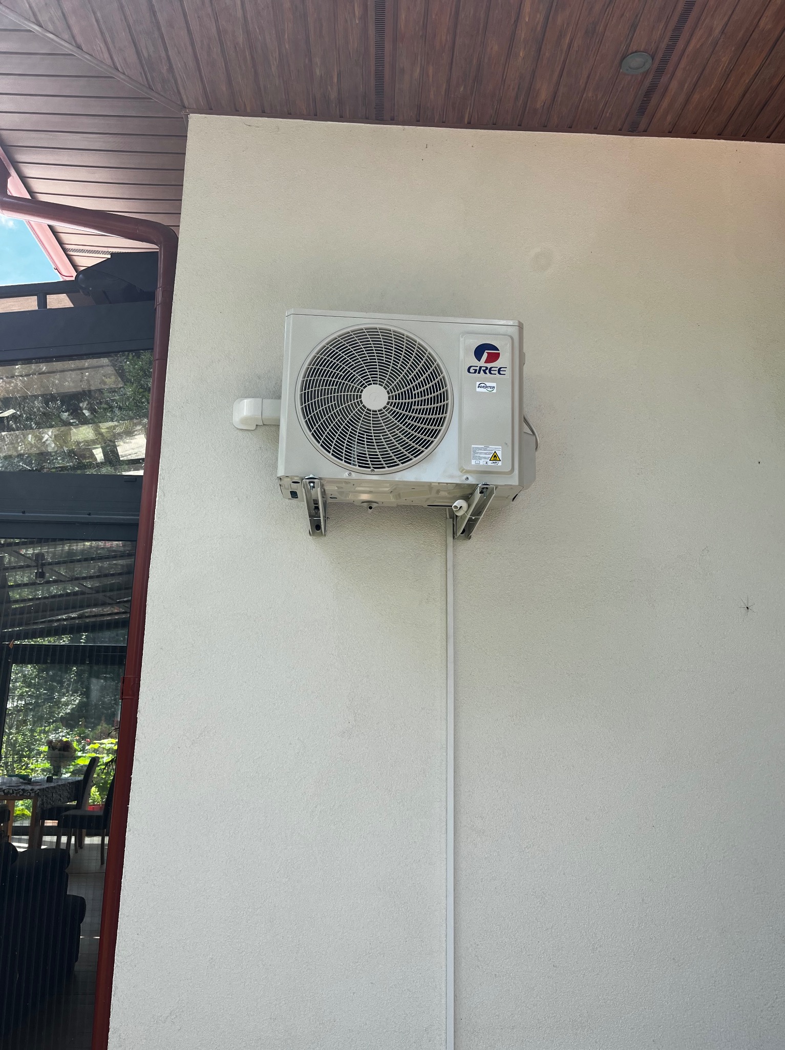 Biała jednostka zewnętrzna klimatyzacji marki Gree zamontowana na białej ścianie budynku mieszkalnego, widoczne metalowe wsporniki i rurka odprowadzająca skropliny.