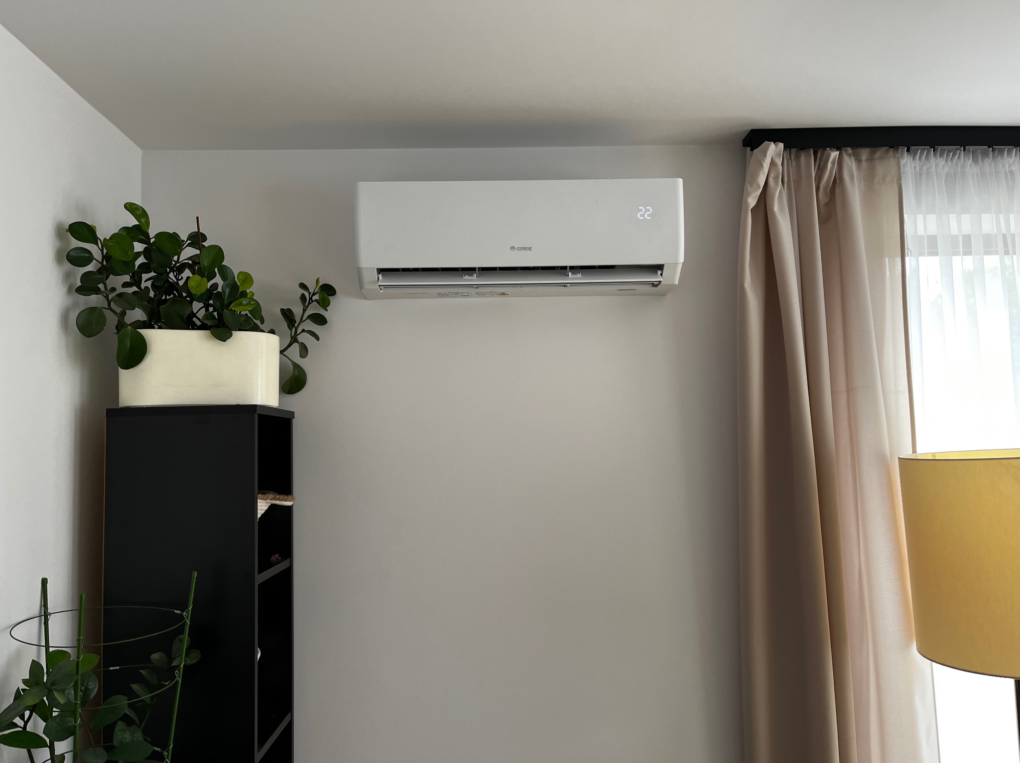 Biały klimatyzator marki Gree zamontowany na ścianie w jasnym wnętrzu, obok czarnego regału z rośliną w białej doniczce i zasłoniętego okna z beżowymi zasłonami.