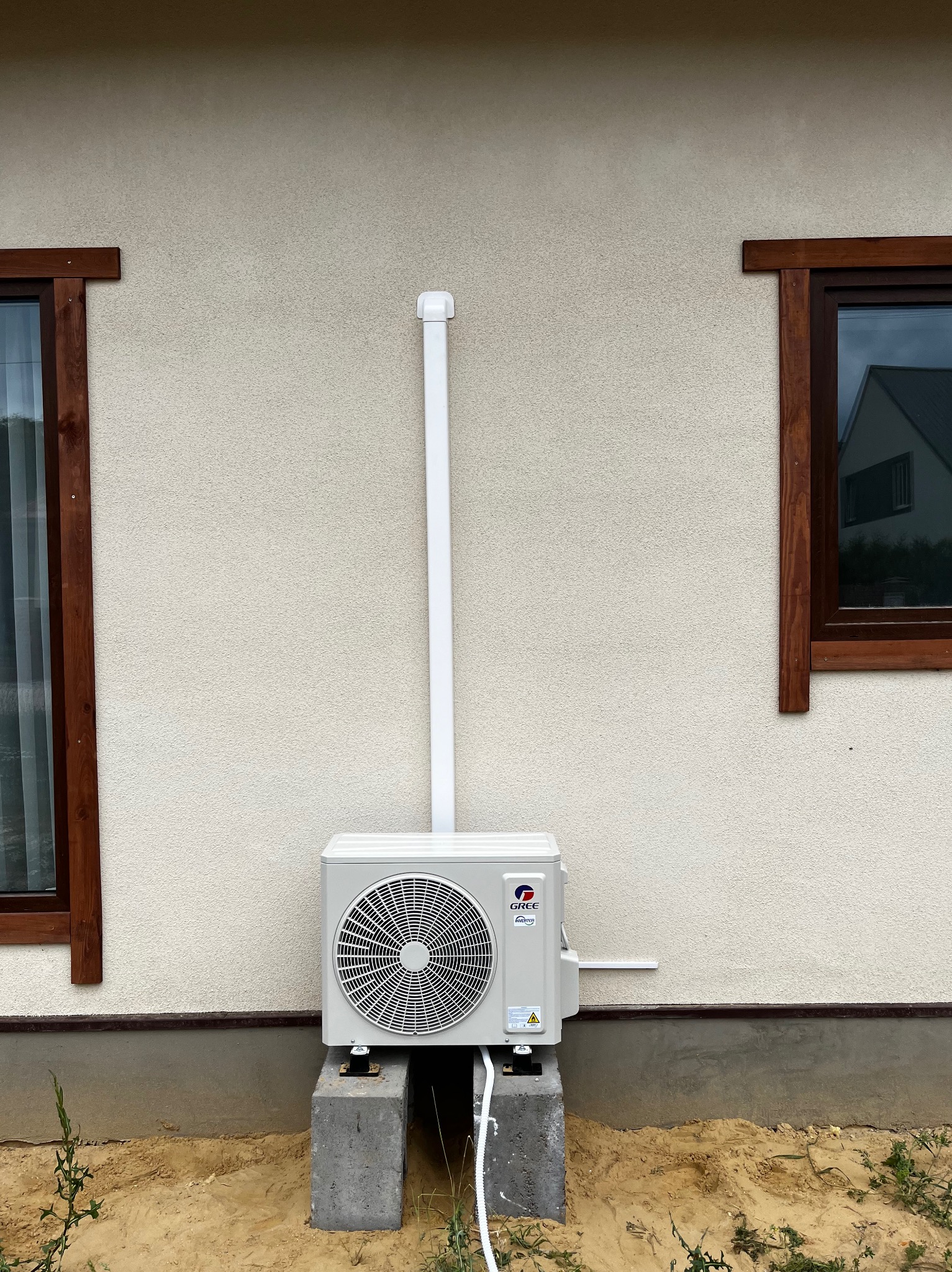 Zewnętrzna jednostka klimatyzacji marki Gree zamontowana na betonowych bloczkach przy ścianie budynku z drewnianą stolarką okienną i pionową rurą odprowadzającą skropliny.