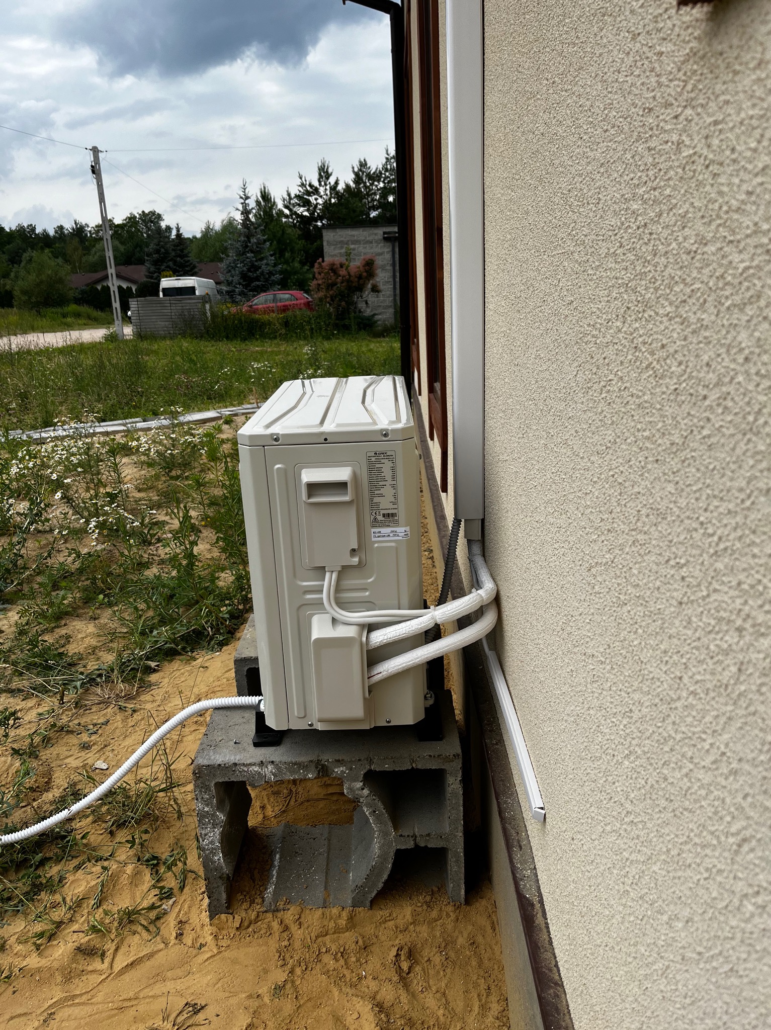 Zewnętrzna jednostka klimatyzacji zamontowana na betonowym fundamencie przy ścianie domu z elewacją typu baranek, widoczne rury odprowadzające czynniki chłodnicze w białej osłonie.