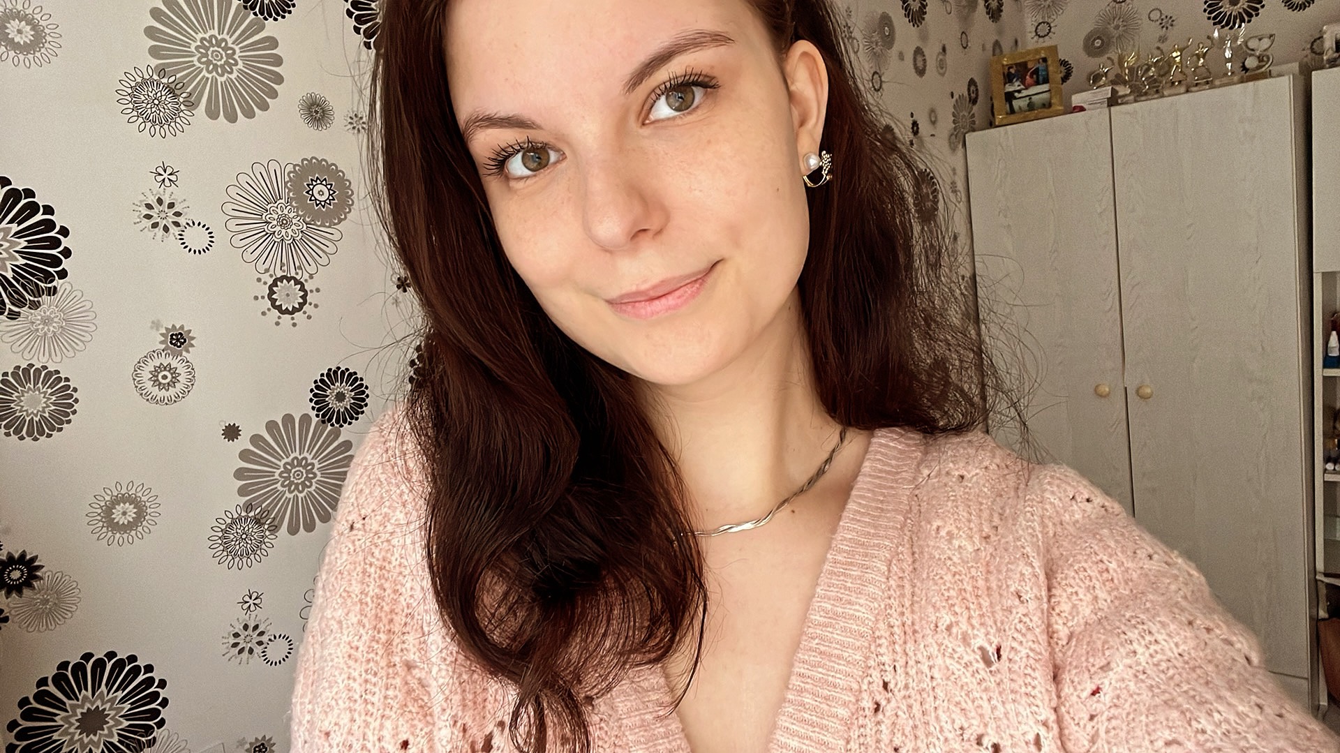 Portret uśmiechniętej młodej kobiety z brązowymi włosami, w różowym swetrze, z delikatną srebrną biżuterią na tle ściany w kwiaty.