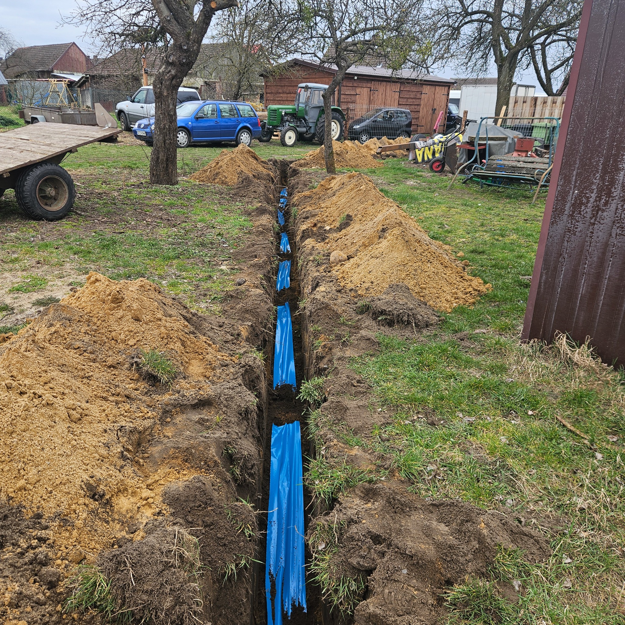 Wykopany rów z widoczną niebieską folią zabezpieczającą, obok hałdy ziemi, na trawiastym terenie z budynkami gospodarczymi i pojazdami w tle.