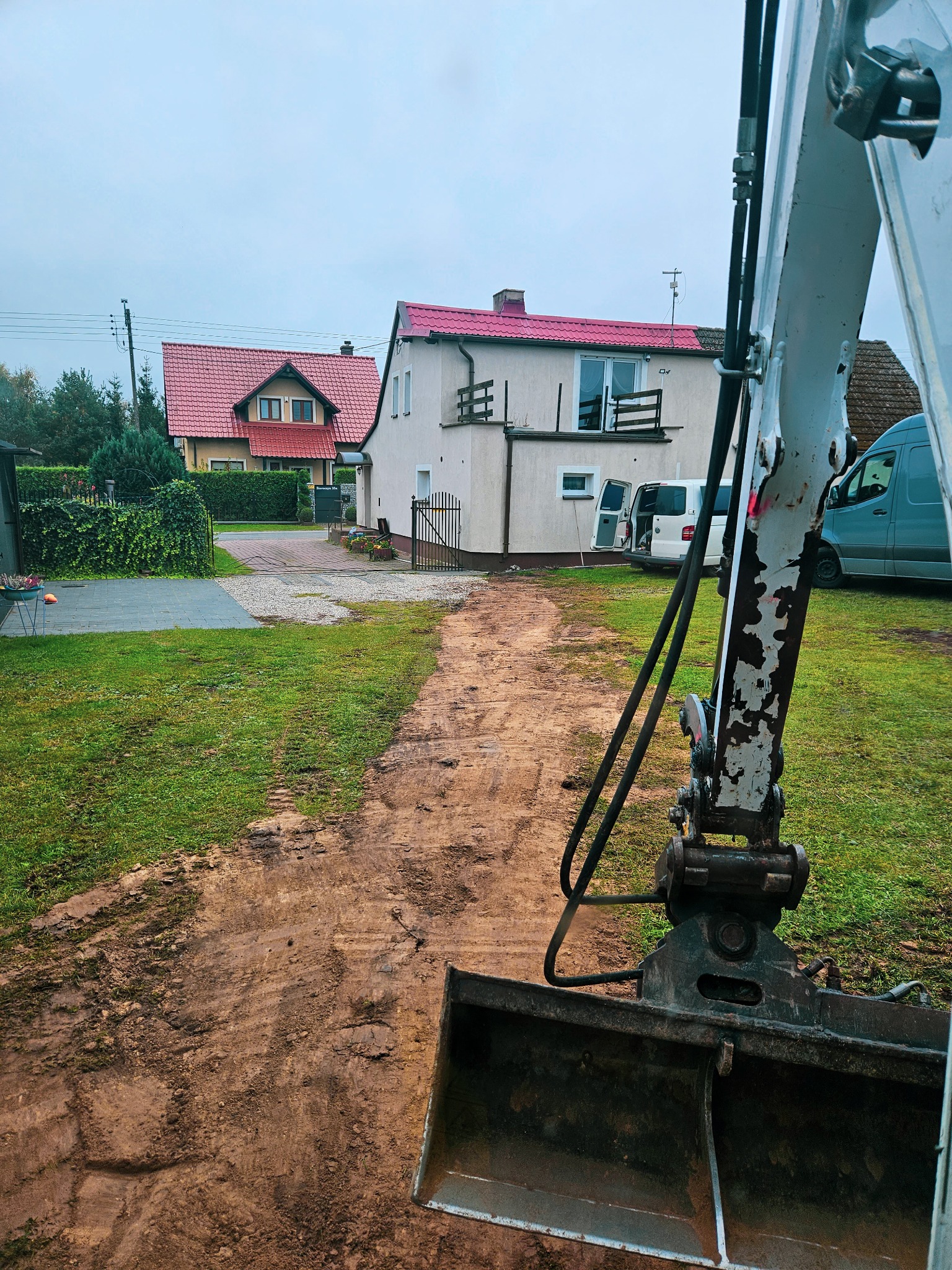 Widok z perspektywy operatora koparki na wykopany pas ziemi w ogrodzie, z dwoma domami w tle i zaparkowanymi samochodami dostawczymi.