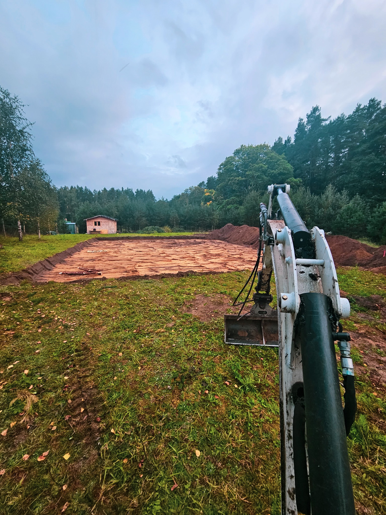 Przygotowany teren pod budowę z widocznym wykopem i mini koparką z przodu, w tle drewniany budynek i drzewa.