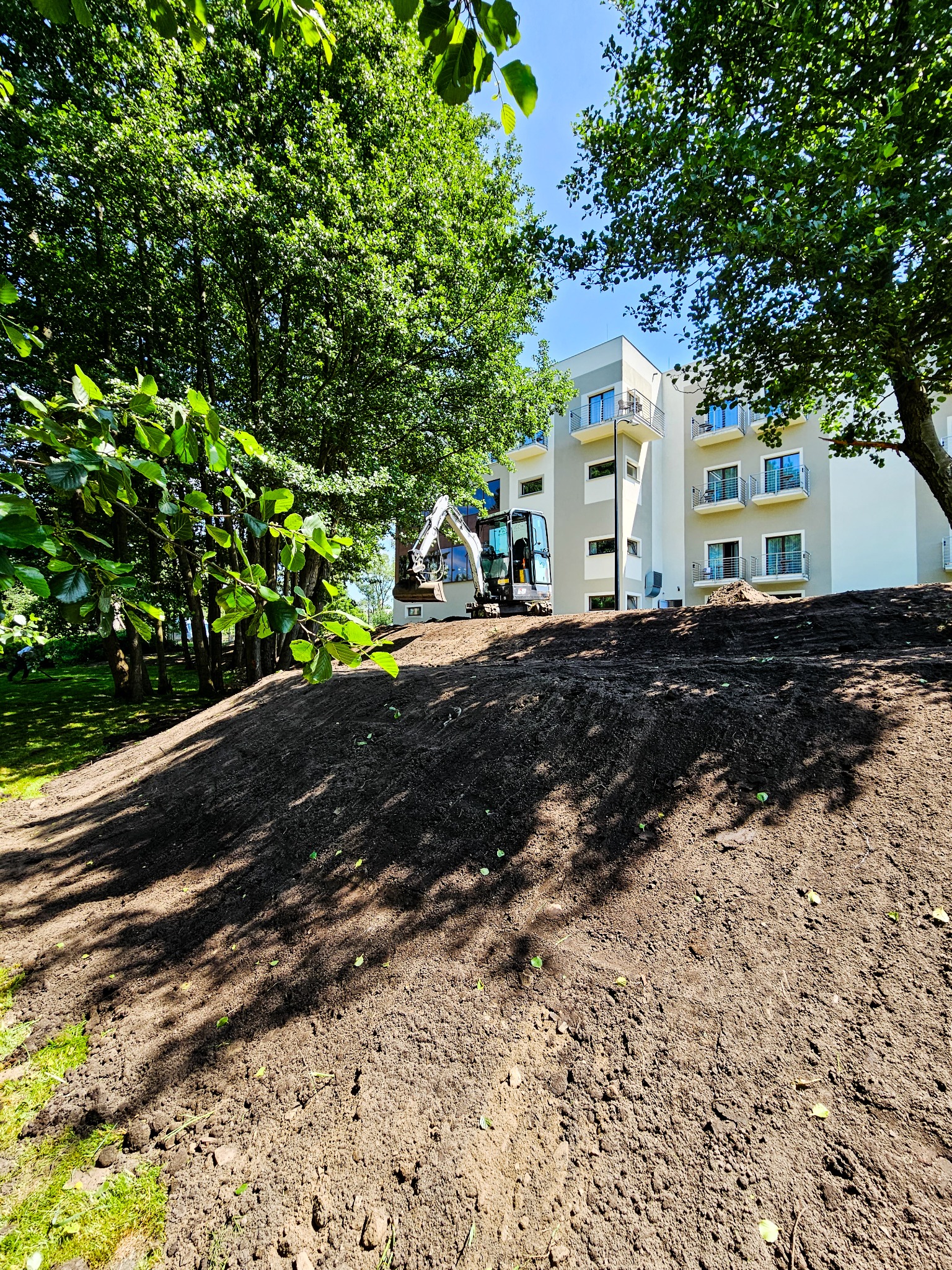 Mini koparka na wzniesieniu z przygotowaną ziemią, w tle budynek apartamentowy i drzewa w słoneczny dzień.