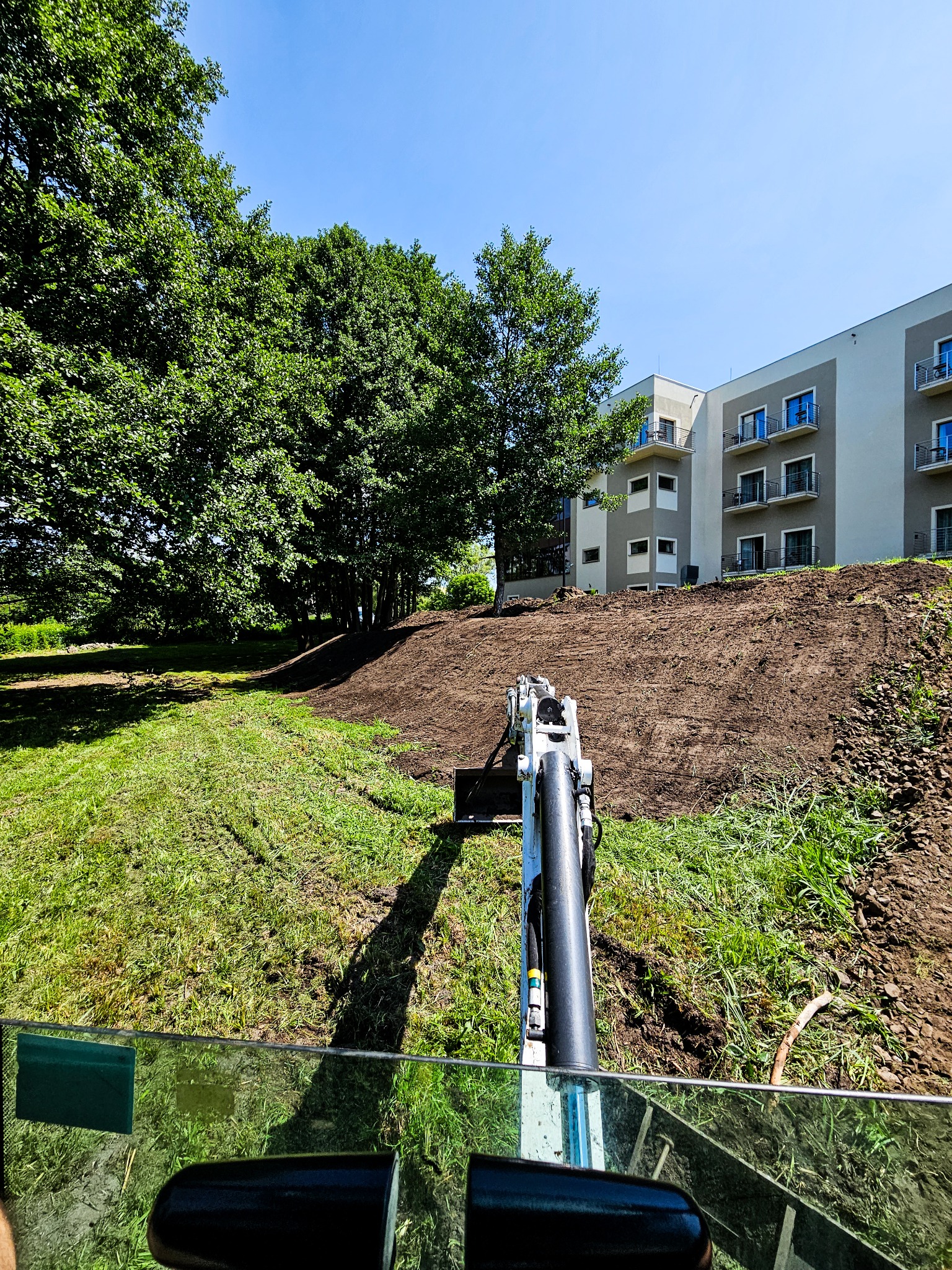 Widok z kabiny operatora na małą koparkę wyrównującą teren pod budynkiem mieszkalnym w słoneczny dzień.