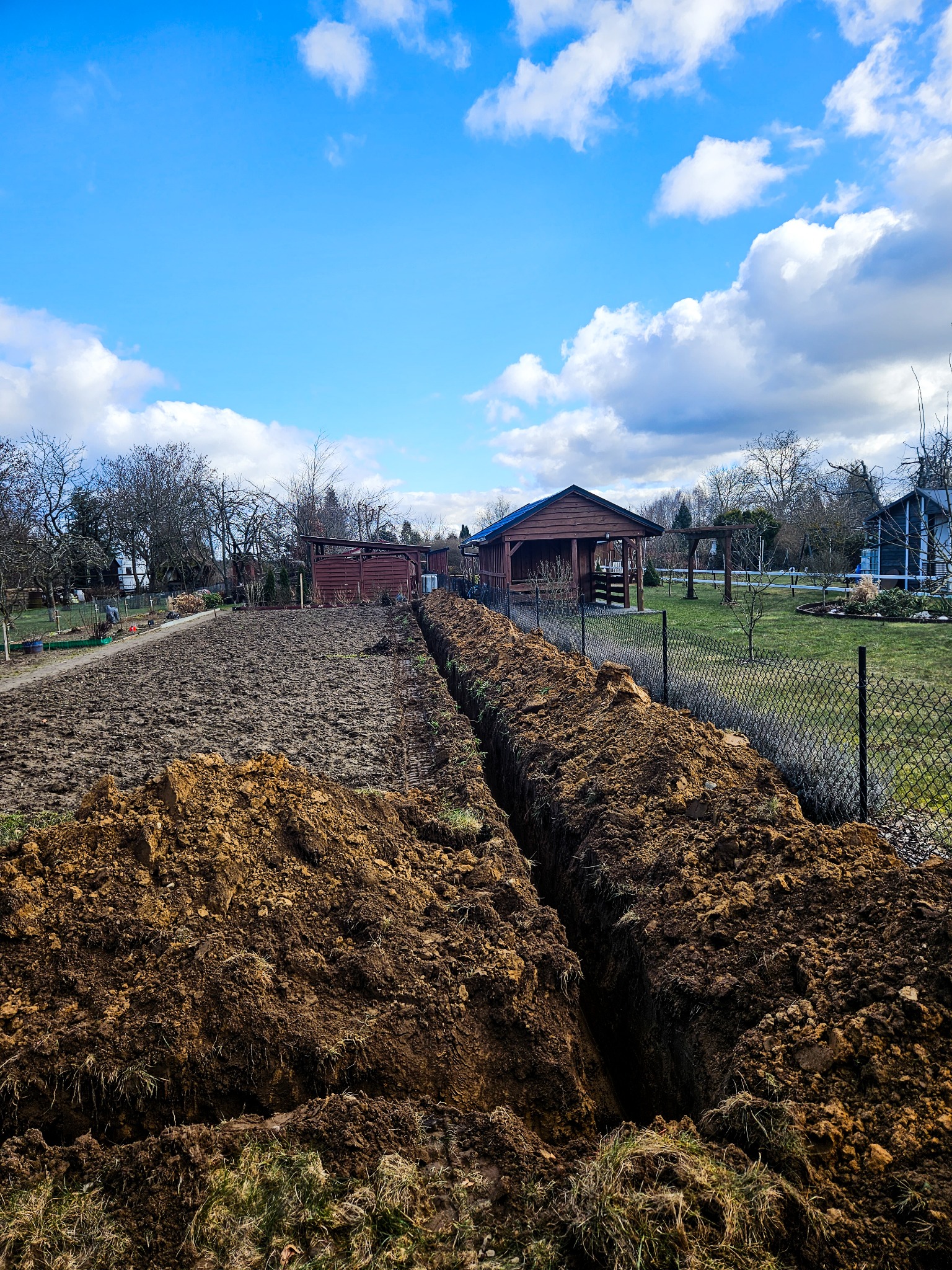 Długi, wąski wykop w ziemi biegnący przez ogród w kierunku drewnianej altany, widoczne świeżo wykopane pryzmy ziemi i fragment siatki ogrodzeniowej.