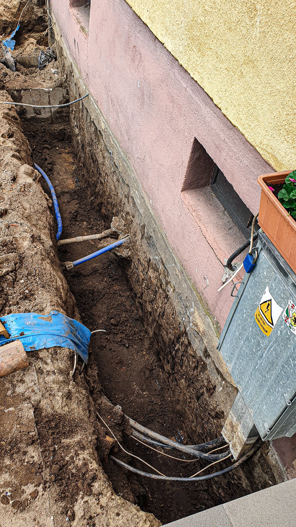 Wykop wzdłuż fundamentu budynku z widocznymi kablami elektrycznymi i rurami instalacyjnymi, skrzynka elektryczna przy ścianie, doniczka z kwiatami na skrzynce.